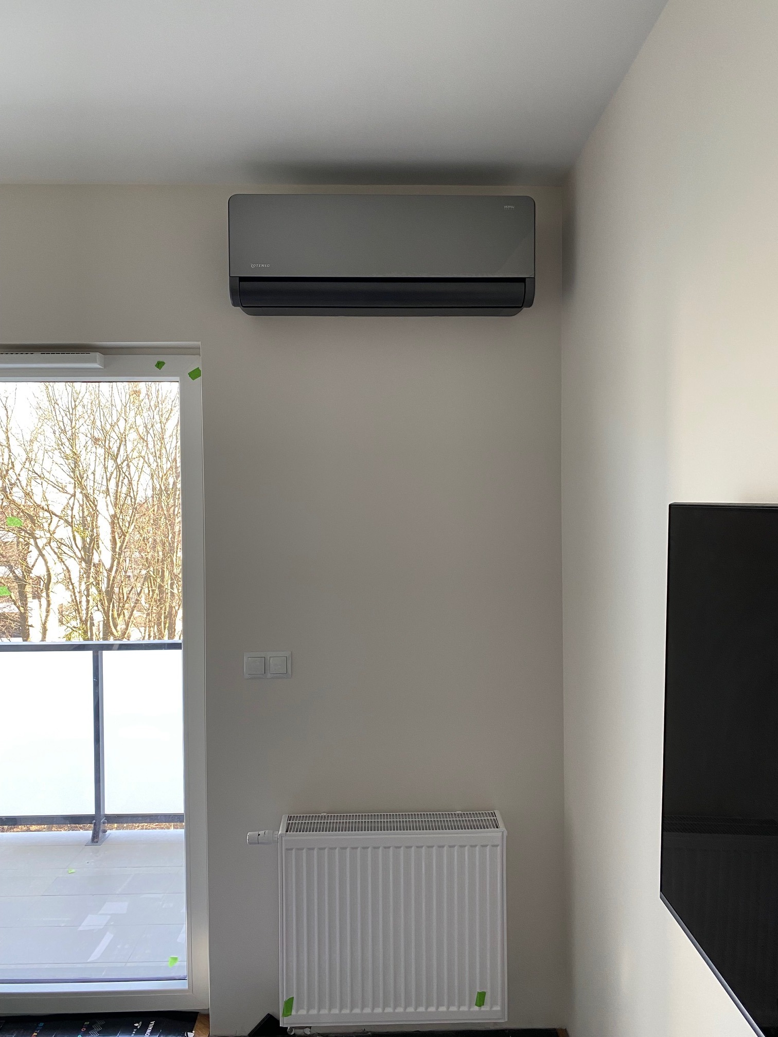 Szary klimatyzator ścienny zamontowany w jasnym pomieszczeniu z grzejnikiem i wyjściem na balkon, widoczne ślady po taśmie malarskiej.