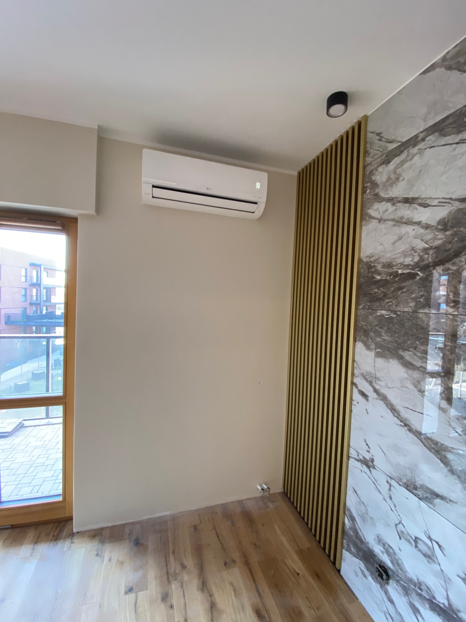 Biały klimatyzator ścienny LG w nowoczesnym wnętrzu z drewnianą podłogą, okno z widokiem na budynek mieszkalny, dekoracyjna ściana z paneli i marmuru.