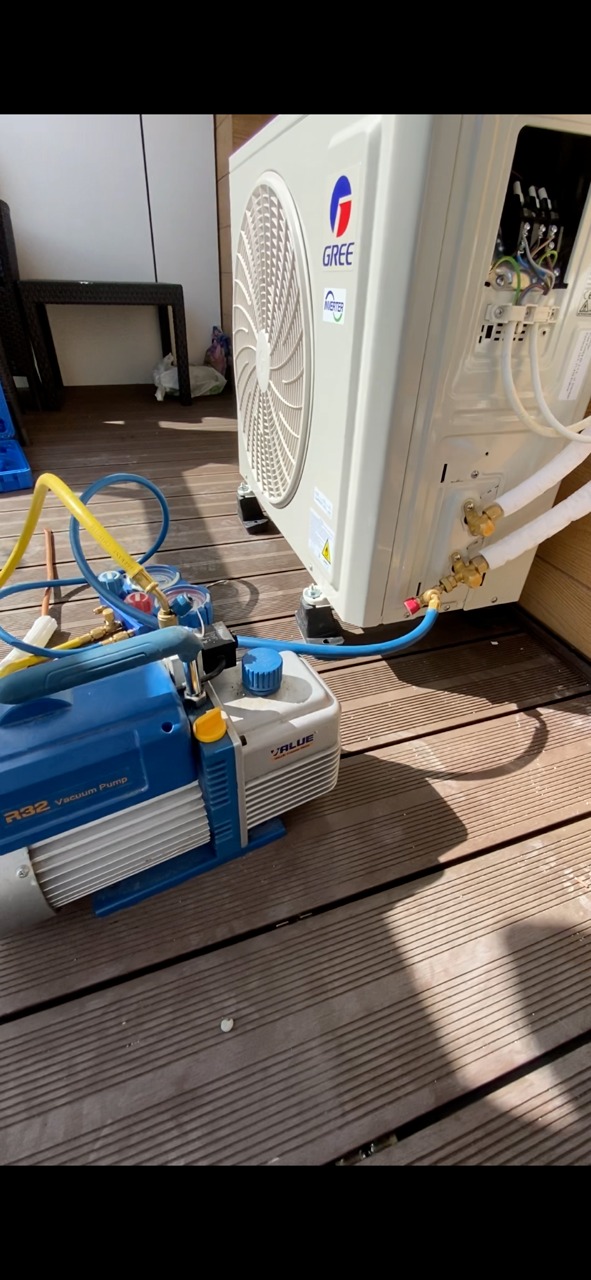 Klimatyzator Gree podczas serwisu na drewnianym tarasie, podłączona niebieska pompa próżniowa Value R32, widoczne przewody chłodnicze i otwarty panel elektryczny klimatyzatora.