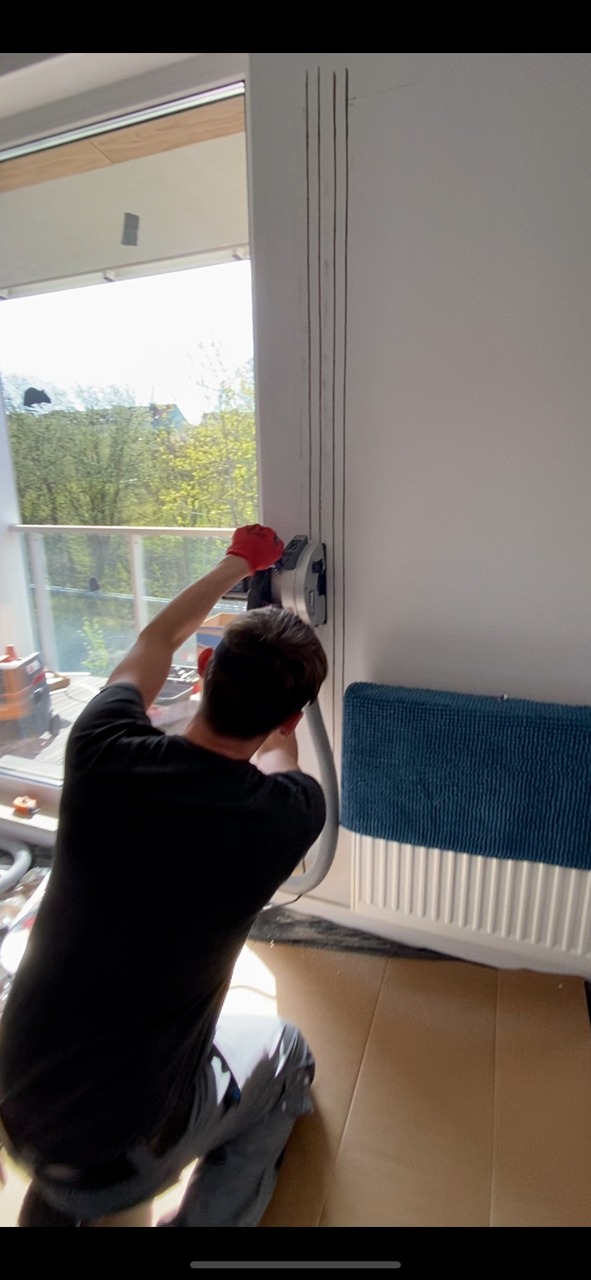 Instalator w czerwonych rękawicach wycina bruzdy w ścianie przy oknie, przygotowując miejsce na rurki, prawdopodobnie pod klimatyzację, z użyciem bruzdownicy z odkurzaczem, radiator osłonięty...