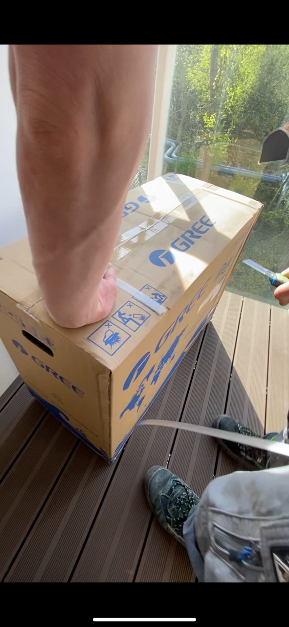 Otwieranie nożem kartonu klimatyzatora marki Gree na zewnątrz, widoczna noga przytrzymująca karton.