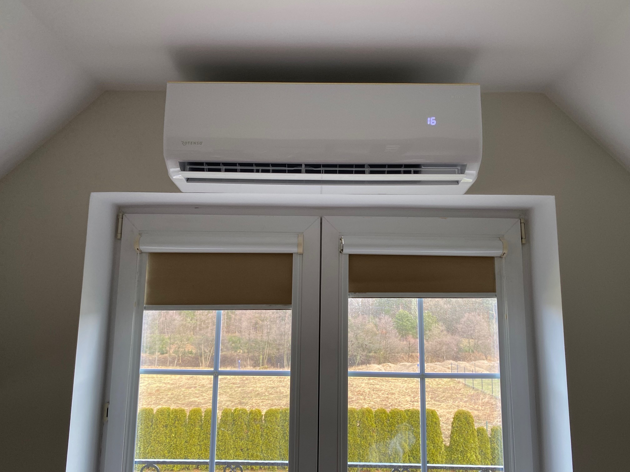 Biały klimatyzator Rotenso zamontowany nad oknem z brązowymi roletami w pokoju na poddaszu, na wyświetlaczu widoczna temperatura 16 stopni Celsjusza.