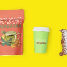 Opakowanie herbaty z grafiką, kubek z napisem 'WOW! Herbatka' i croissant posypany cukrem pudrem na żółtym tle.