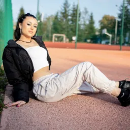 Młoda kobieta w sportowym stroju pozuje siedząc na tle boiska sportowego z zielonym ogrodzeniem.