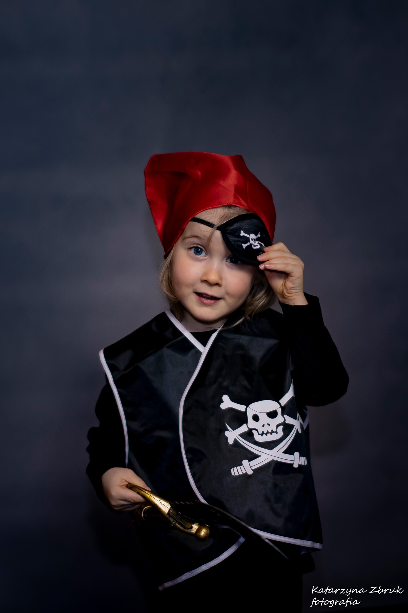 Uśmiechnięta blond dziewczynka w stroju pirata z czerwoną bandaną i opaską na oku z symbolem czaszki, trzymająca złotą szablę na ciemnoszarym tle.