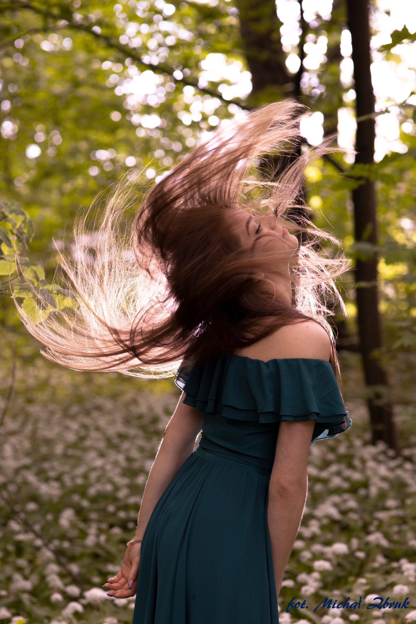 Kobieta w zielonej sukience odwraca głowę, a jej włosy falują w powietrzu na tle kwitnącego lasu.