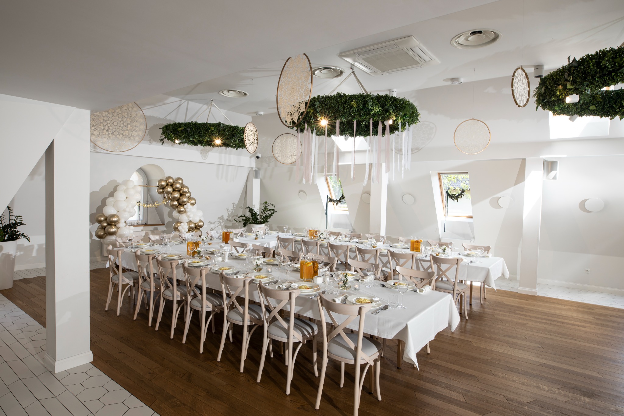 Uroczysta sala z długimi stołami nakrytymi białymi obrusami, dekoracje z zieleni i okręgów zawieszone pod sufitem, aranżacja z balonów przy oknie, drewniana podłoga, neutralna kolorystyka.