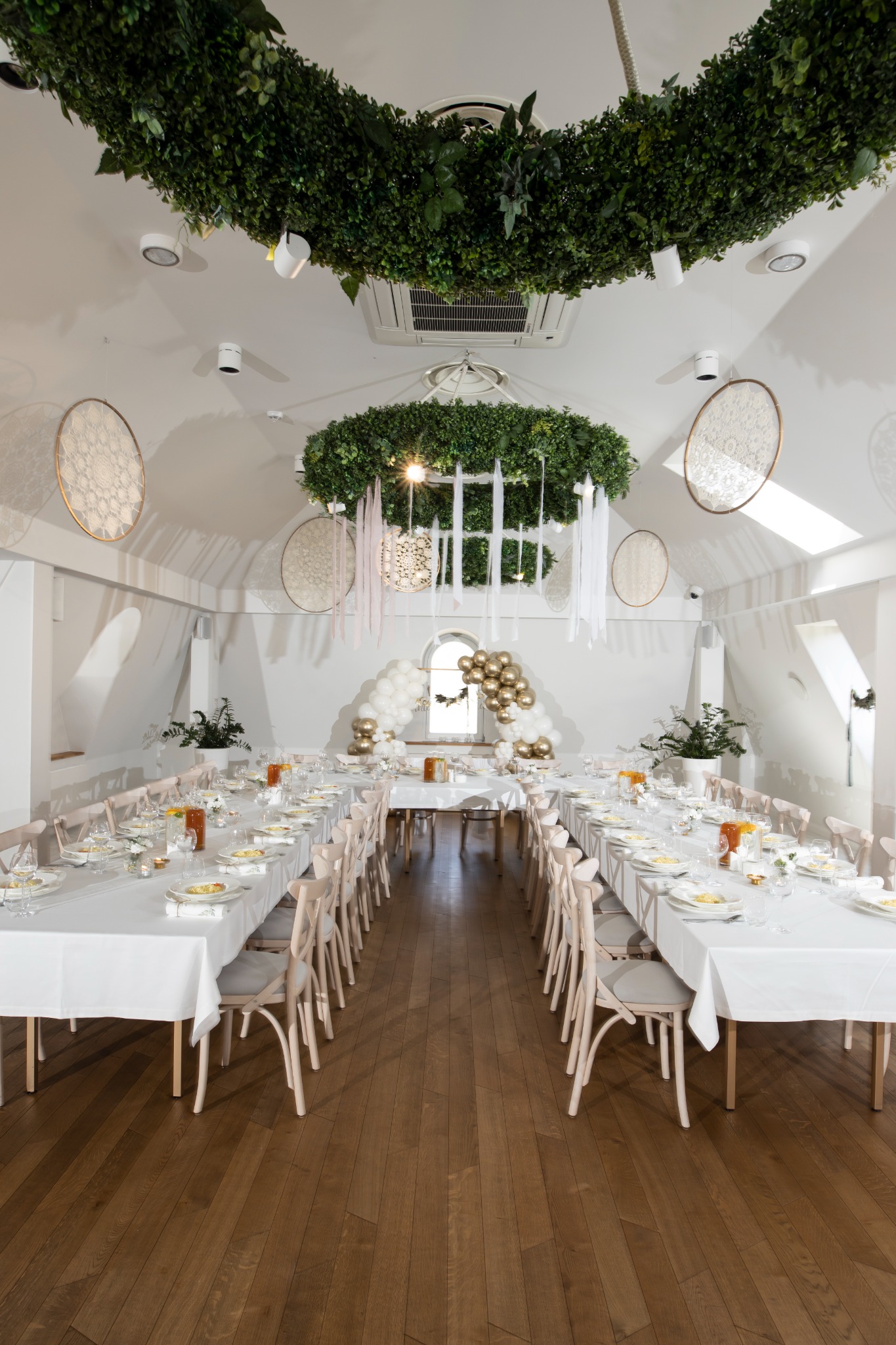 Uroczysta sala w Woli przygotowana na przyjęcie, z długimi stołami nakrytymi białymi obrusami, dekoracjami z zieleni pod sufitem i balonami przy oknie.