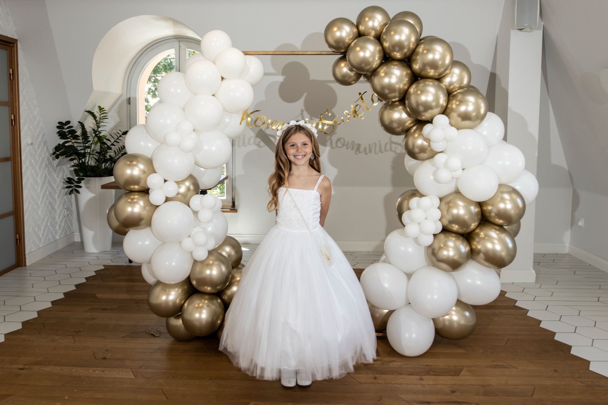 Uśmiechnięta dziewczynka w białej sukience komunijnej, pozująca na tle dekoracji z białych i złotych balonów, z napisem 'Komunia Święta'.
