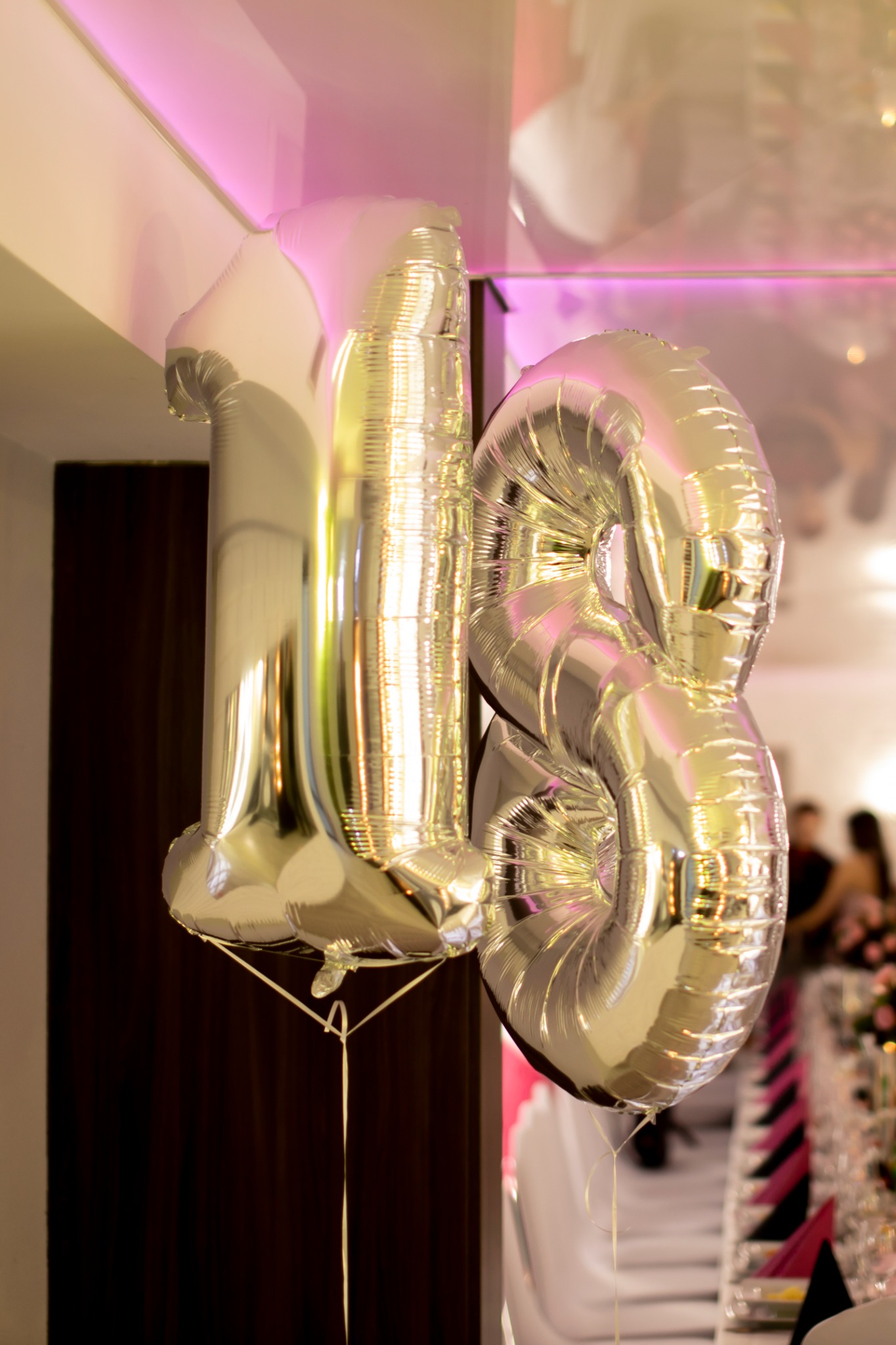 Dwa srebrne balony w kształcie cyfr '1' i '8', zawieszone pod sufitem z dekoracyjnym oświetleniem, w tle widoczny stół z nakryciami i goście.