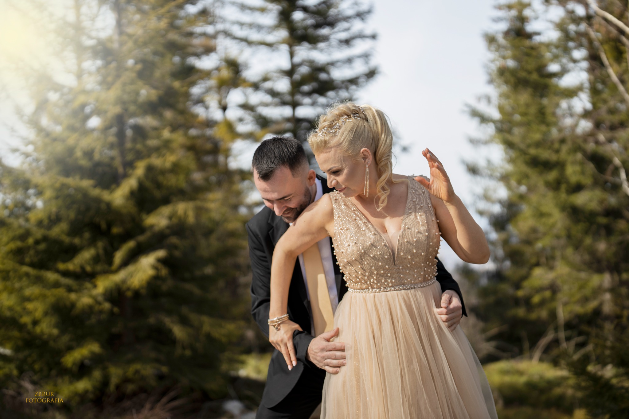 Uśmiechnięta para w plenerze: mężczyzna w czarnym garniturze obejmuje kobietę w beżowej sukni z perłami i tiarą we włosach, na tle lasu iglastego.