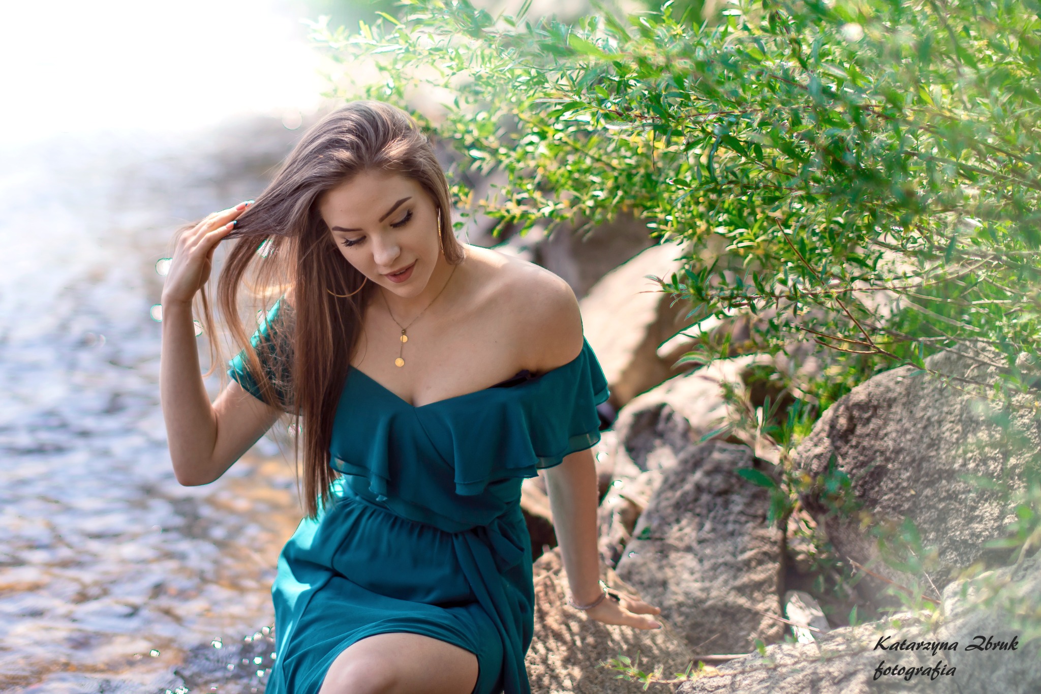 Młoda kobieta w szmaragdowej sukience pozuje nad strumieniem, dotykając włosów na tle zieleni i skał.