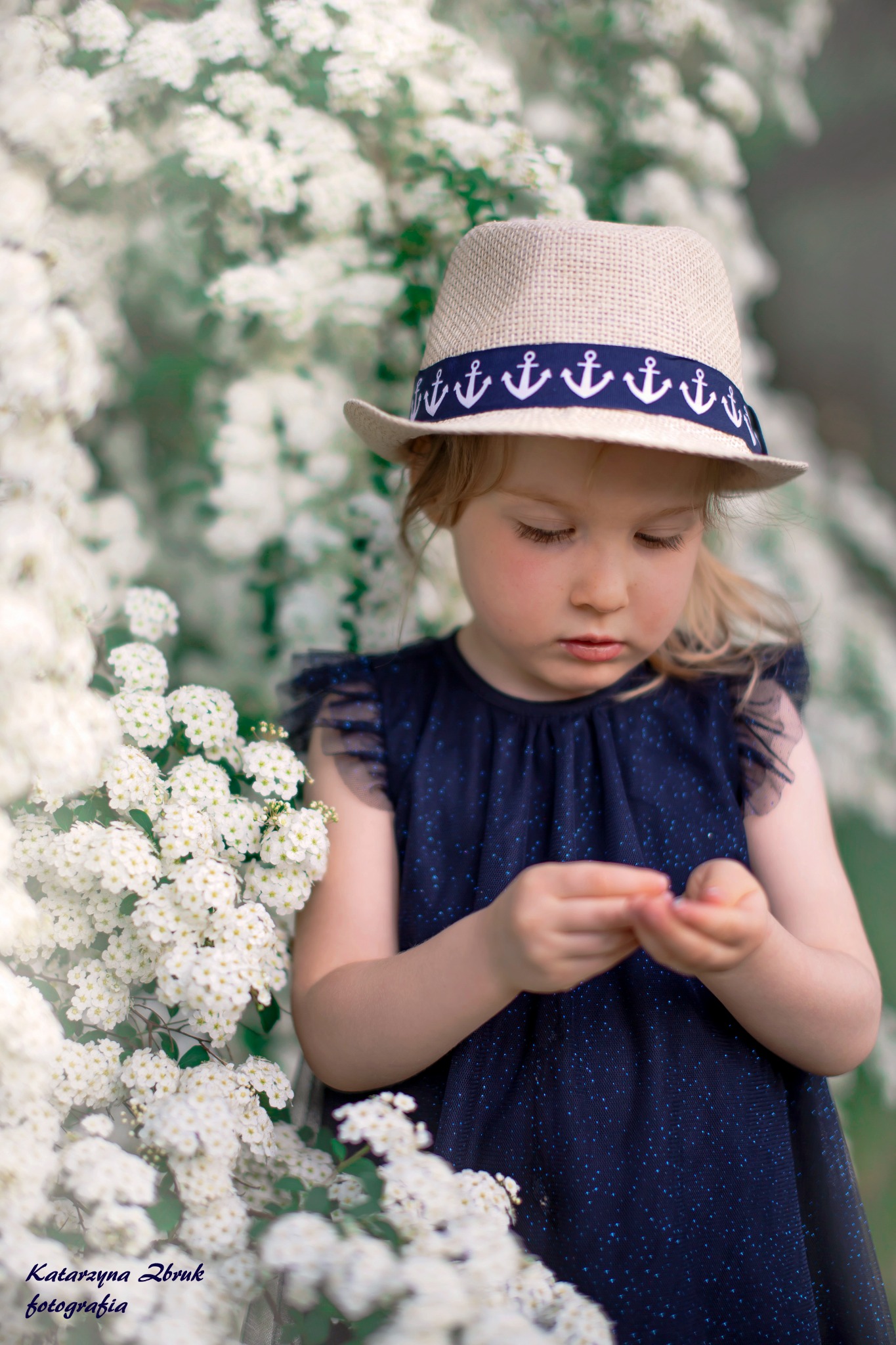 Dziewczynka w słomkowym kapeluszu z motywem kotwic, w granatowej sukience w kropki, bawi się drobnymi kwiatami na tle kwitnącego krzewu tawuły.