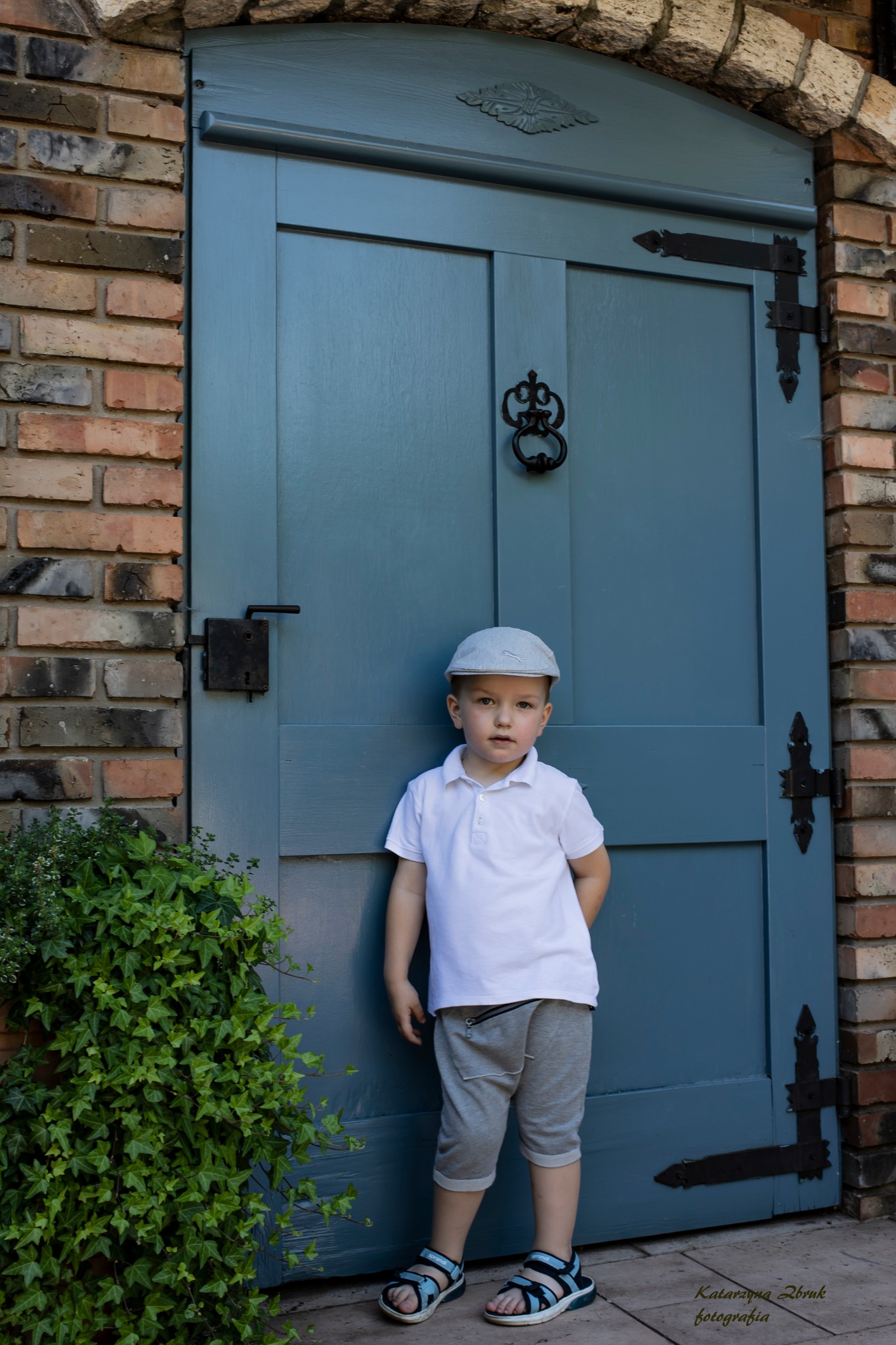 Chłopiec w czapce i sandałach pozuje na tle niebieskich drzwi z dekoracyjnymi okuciami i ceglanej ściany.