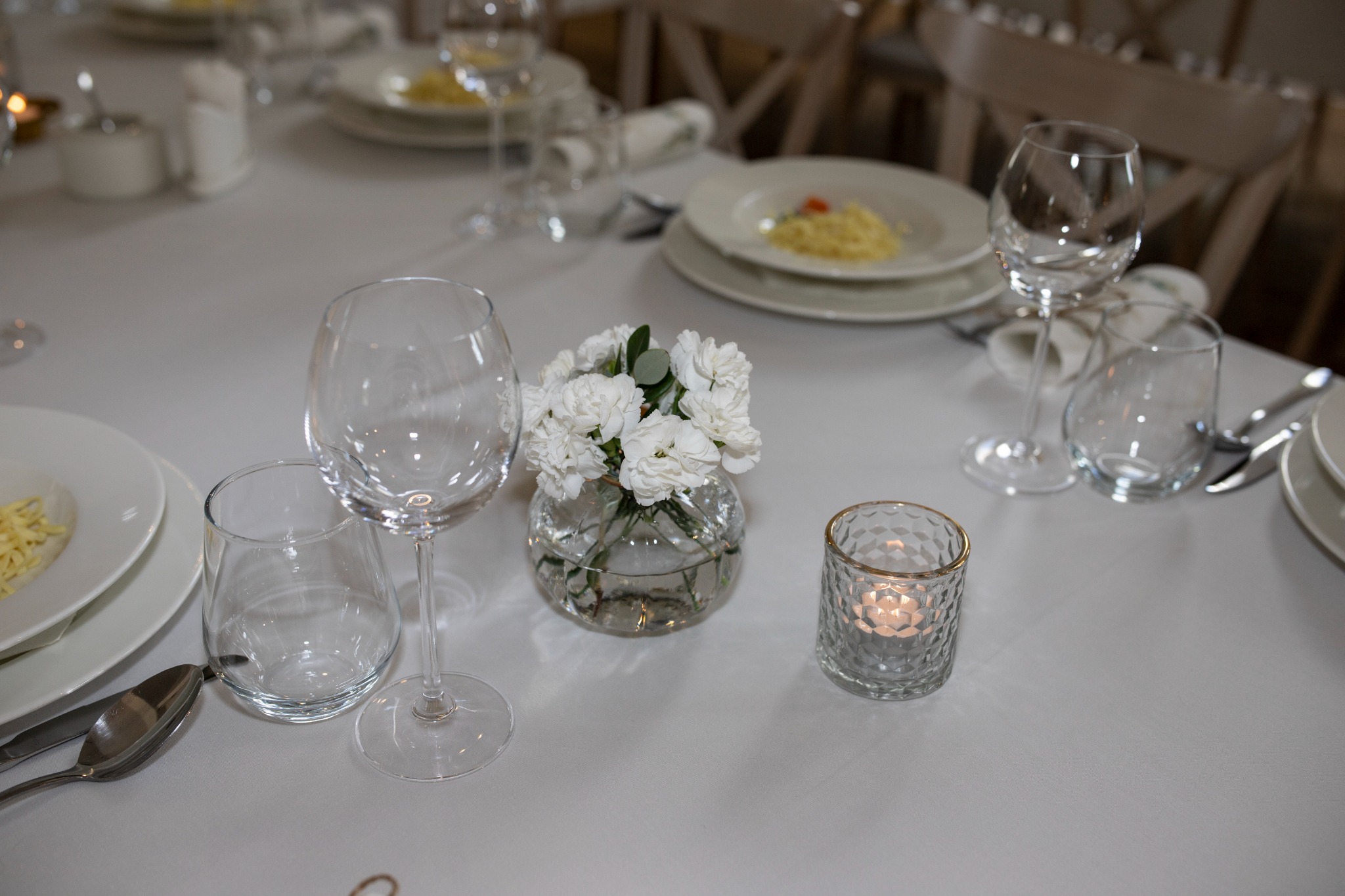Elegancko nakryty stół z białym obrusem, zastawą, szklankami, wazonikiem z białymi goździkami i świecą w ozdobnym szkle, przygotowany do uroczystego obiadu lub kolacji.