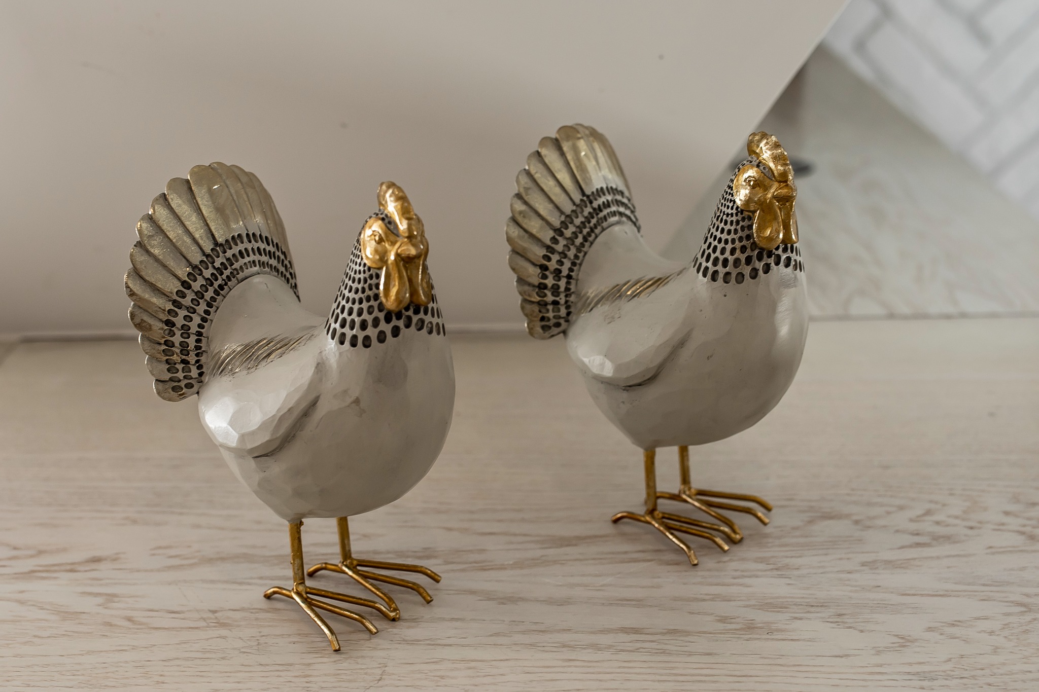 Dwie dekoracyjne figurki kur w kolorze srebrnym z elementami złota, stojące na jasnym blacie.