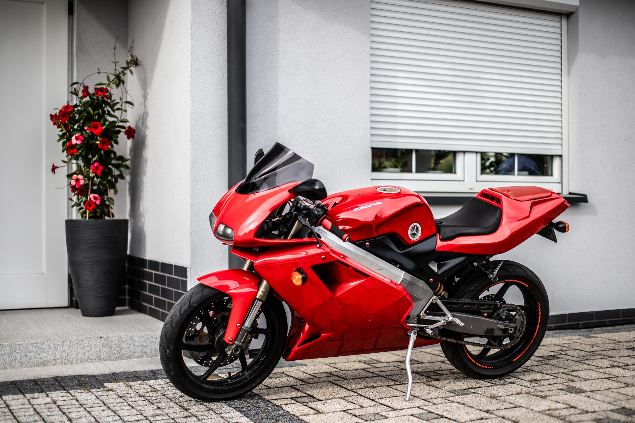 Czerwony motocykl Cagiva Mito na kostce brukowej przed białym budynkiem z zamkniętymi żaluzjami i donicą z czerwonymi kwiatami.
