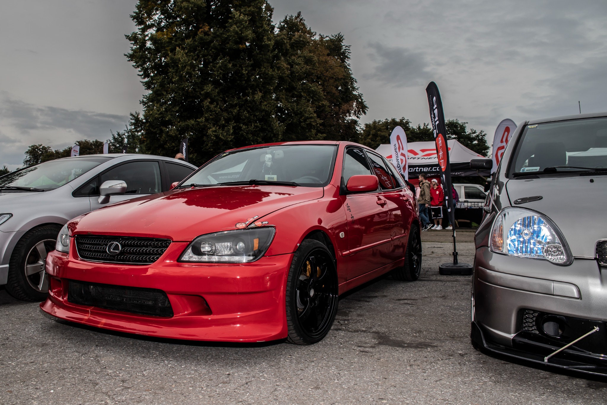Czerwony samochód Lexus IS200/IS300 na zlocie tuningowym, widoczny z przodu, z czarnymi felgami i naklejkami na reflektorze, otoczony innymi pojazdami i stoiskami.