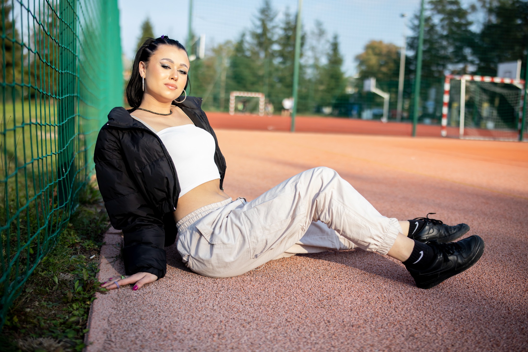 Młoda kobieta w sportowym stroju pozuje siedząc na tle boiska sportowego z zielonym ogrodzeniem.