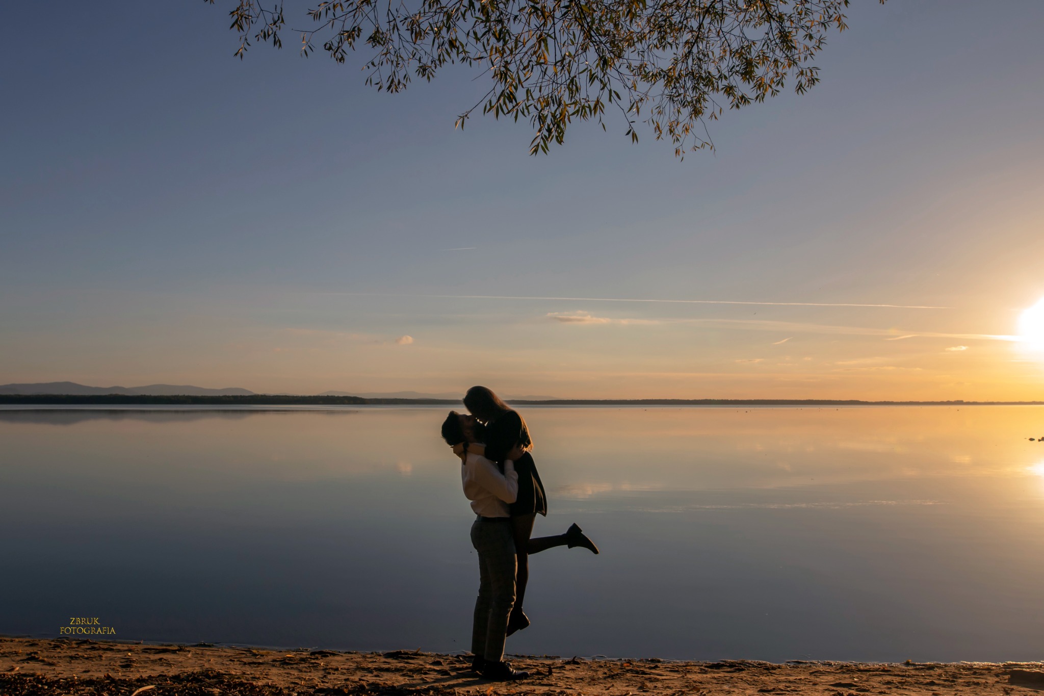 Zakochana para w objęciach na plaży o zachodzie słońca, jezioro w tle odbija niebo, gałęzie drzewa w górnej części kadru.