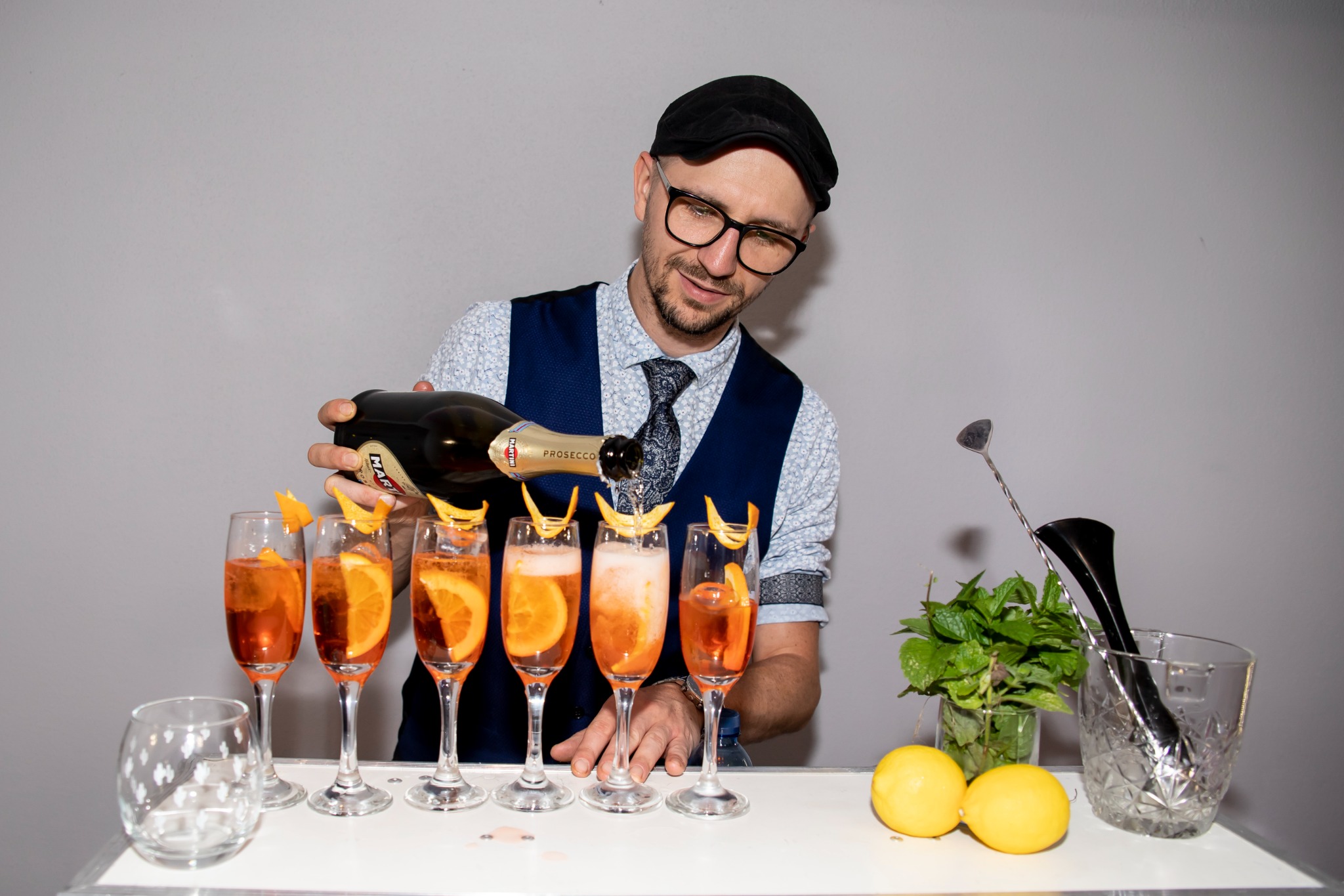 Barmam w czapce wlewa Prosecco Martini do kieliszków z Aperolem i pomarańczą, obok wiaderko z miętą i cytrynami.