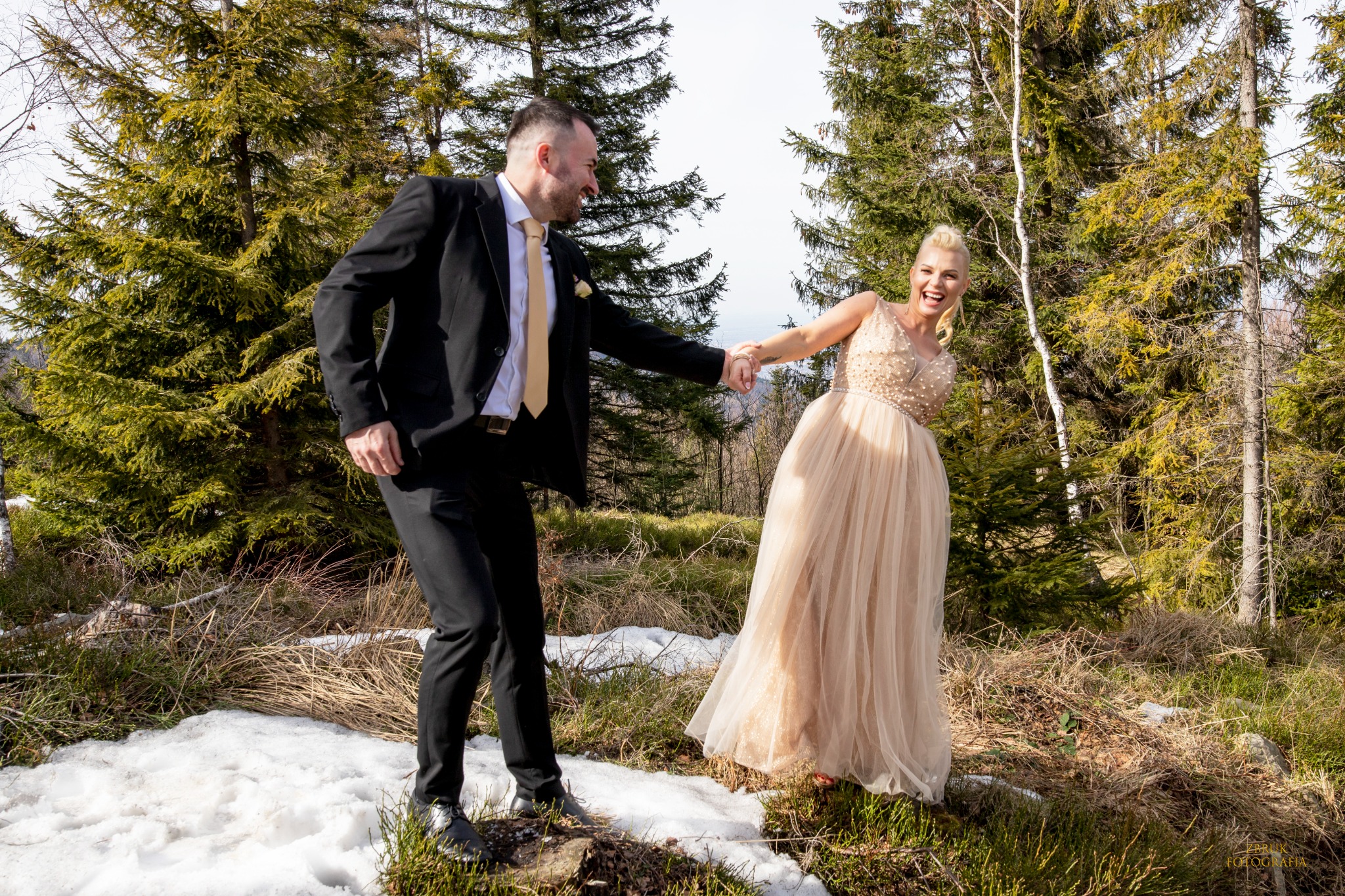 Uśmiechnięta para w strojach ślubnych trzymająca się za ręce na tle leśnego krajobrazu z resztkami śniegu.