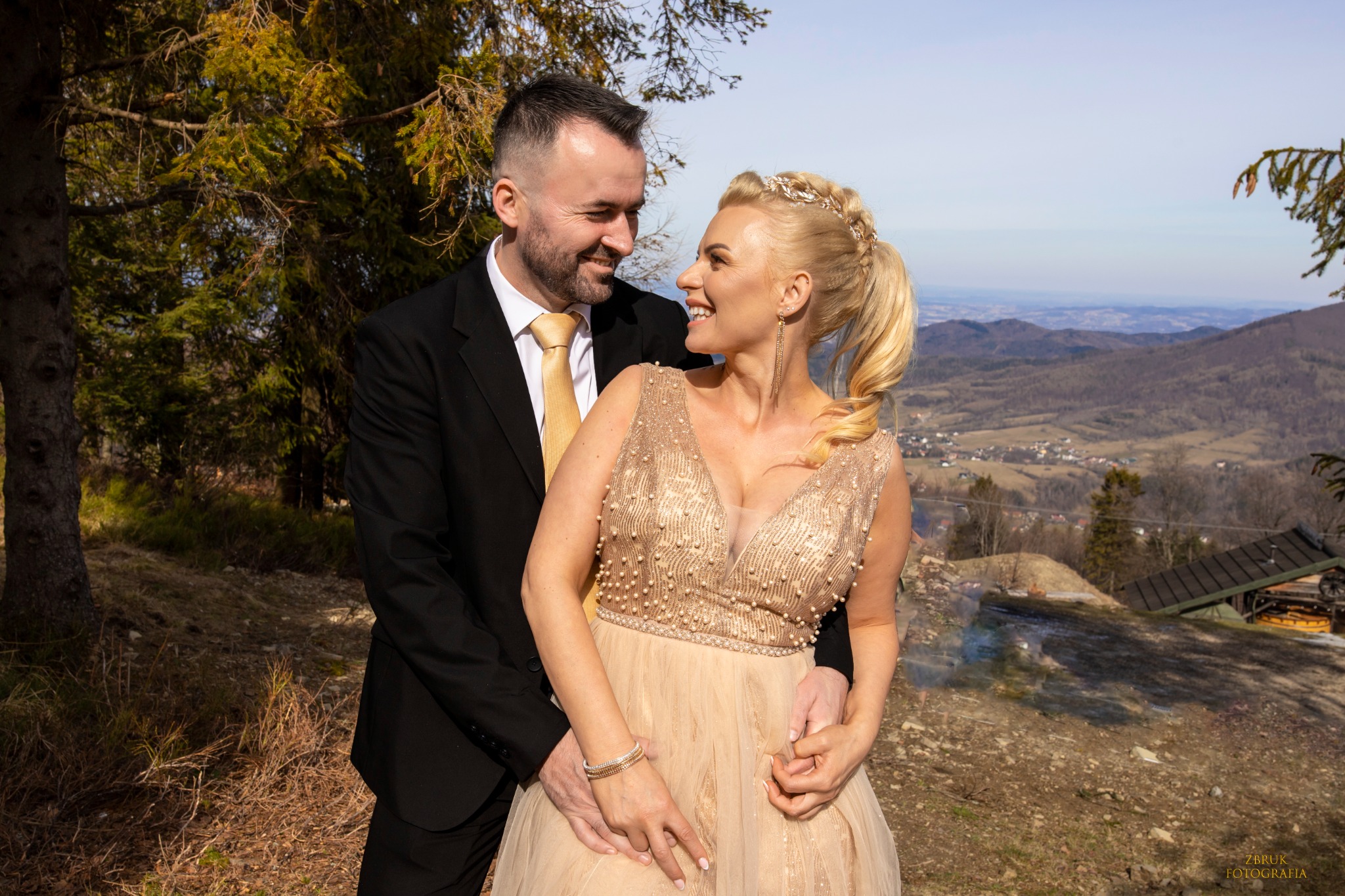 Uśmiechnięta para w strojach ślubnych, on w czarnym garniturze i złotym krawacie, ona w beżowej sukni z perłami, obejmująca się na tle górskiego krajobrazu.