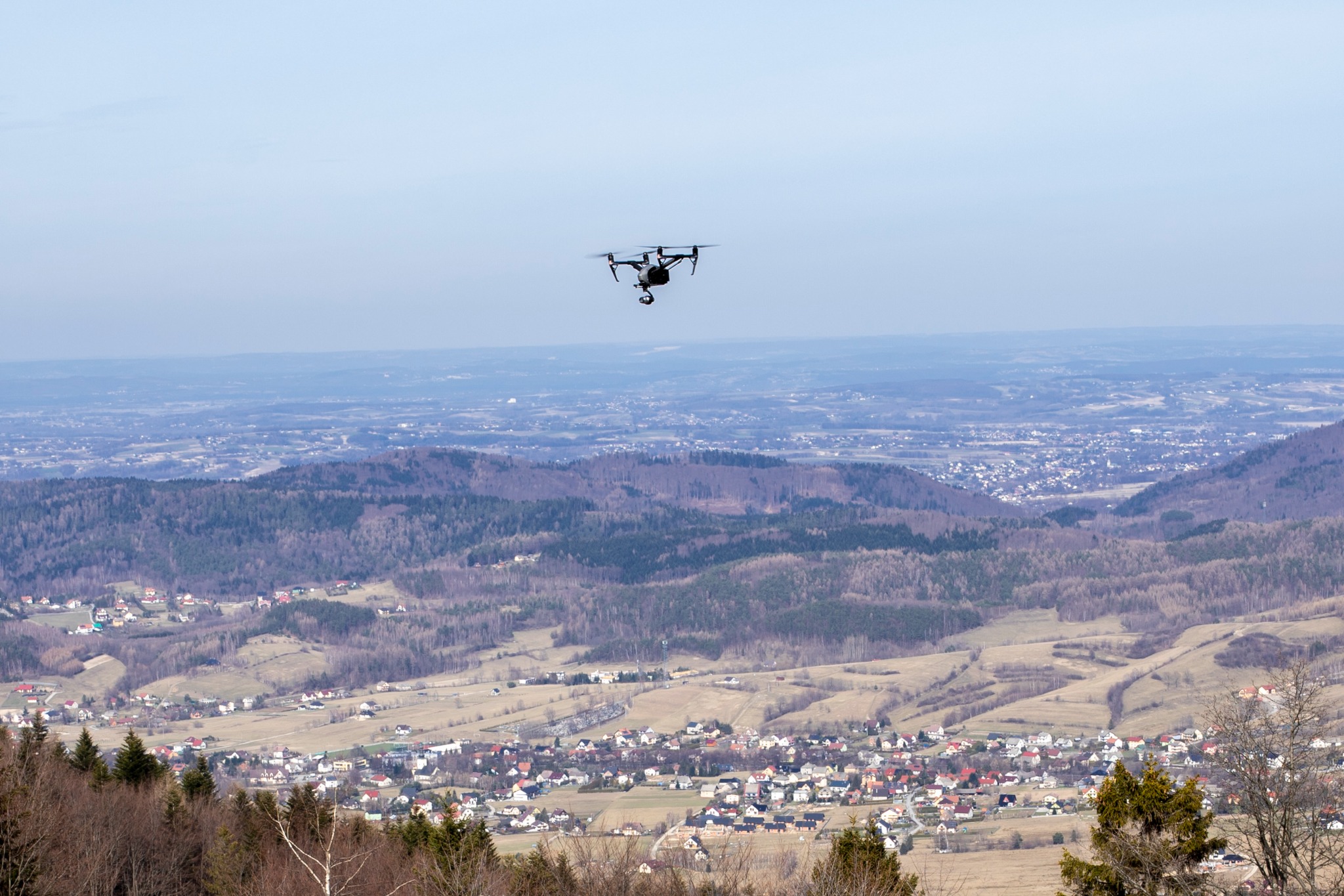 Czarny dron w locie nad pagórkowatym terenem z widoczną zabudową mieszkalną w oddali, sfotografowany w jasny dzień.