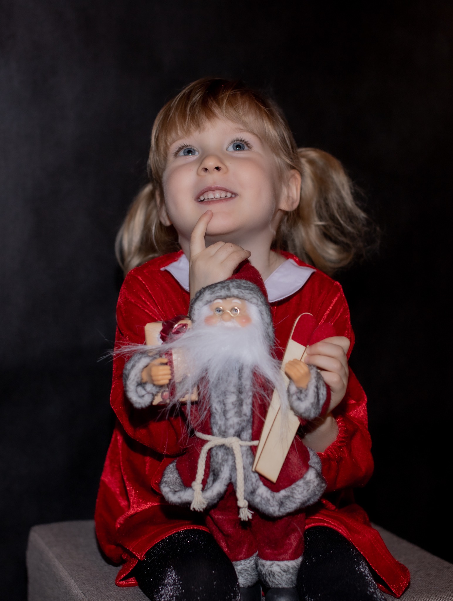 Dziewczynka w czerwonej sukience trzyma pluszowego Mikołaja, spoglądając w górę z zadumą.