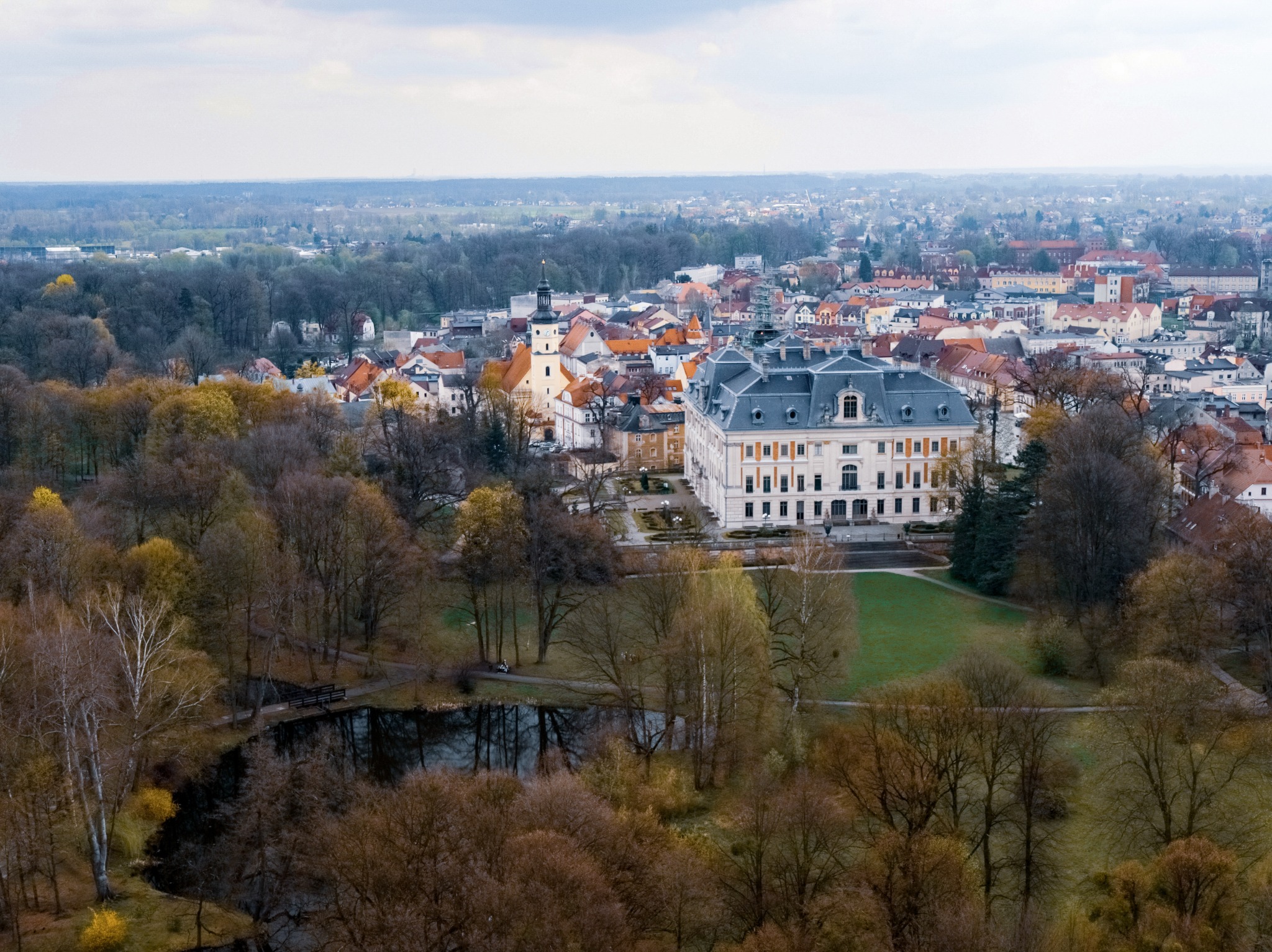 Panorama miasta z perspektywy lotu ptaka, z widocznym barokowym pałacem z szarym dachem, wieżą kościelną, parkiem z jeziorkiem i mostkiem oraz zabudową miejską w tle.