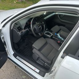 RichMont Service - Wnętrze białego samochodu Audi A4 S-Line z otwartymi drzwiami, widoczne czarne dywaniki i szara tapicerka foteli, butelka wody na konsoli środkowej.