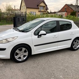 RichMont Service - Biały Peugeot 207 zaparkowany na szarym asfalcie, z widocznymi domami w tle i zielenią, prezentowany z boku w pochmurny dzień.