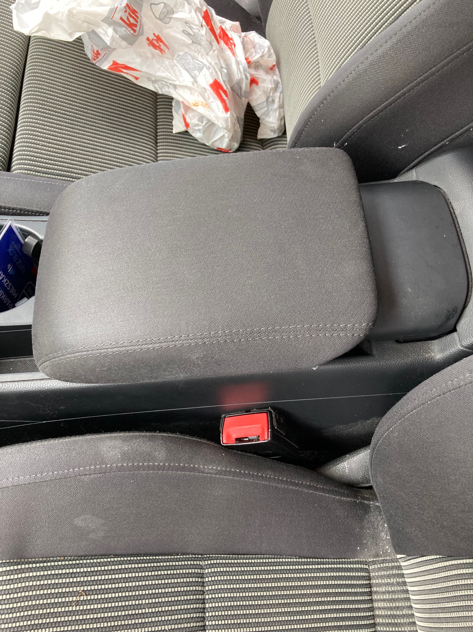 Wnętrze samochodu z widocznymi fotelami z szarej tapicerki z plamami i okruchami, podłokietnikiem oraz torbą reklamową na siedzeniu