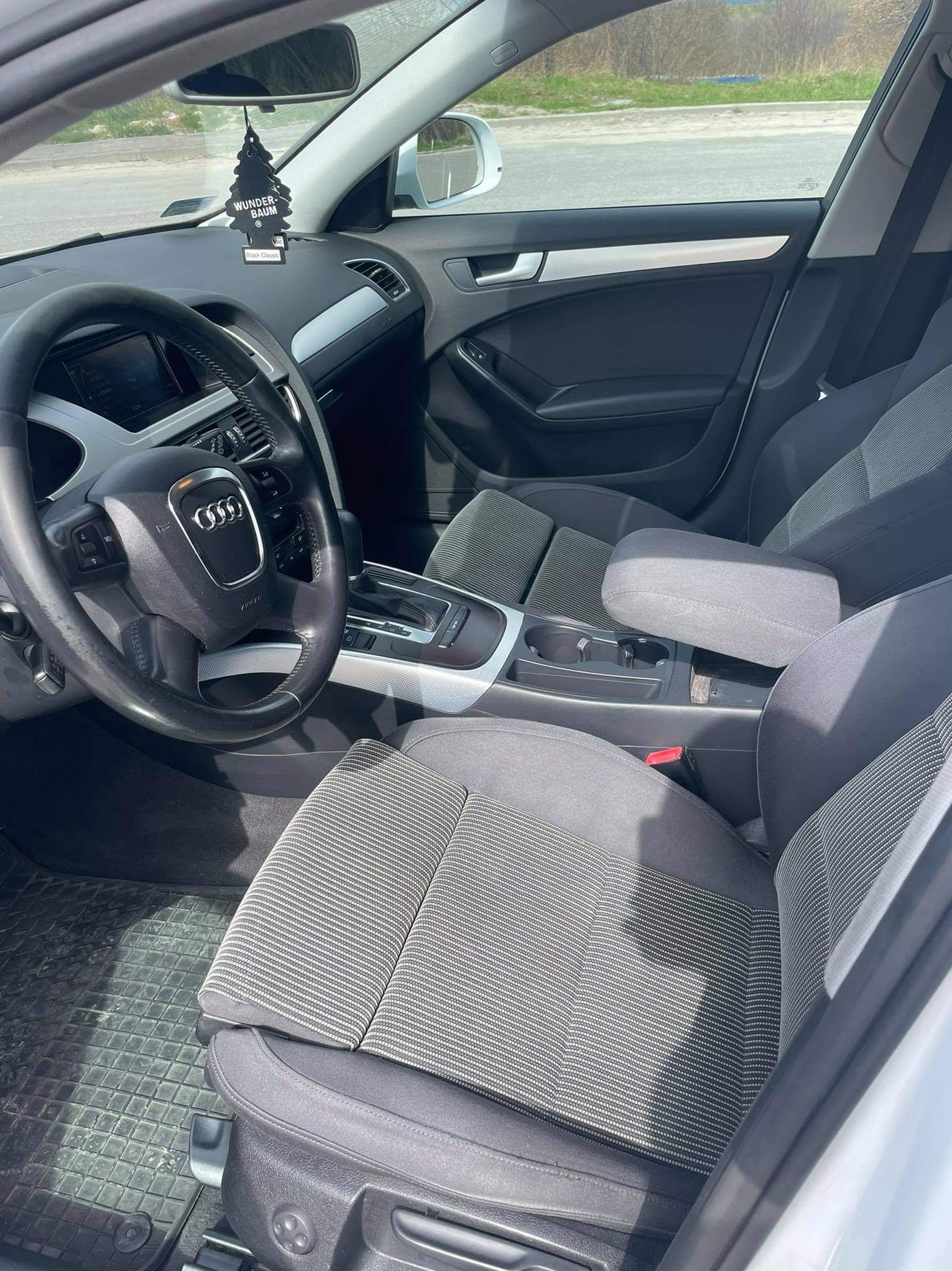 Wnętrze samochodu Audi A4 z szarą tapicerką, widoczna kierownica, deska rozdzielcza z zawieszką zapachową Wunder-Baum Black Classic, fotele z materiałową fakturą i gumowe dywaniki.