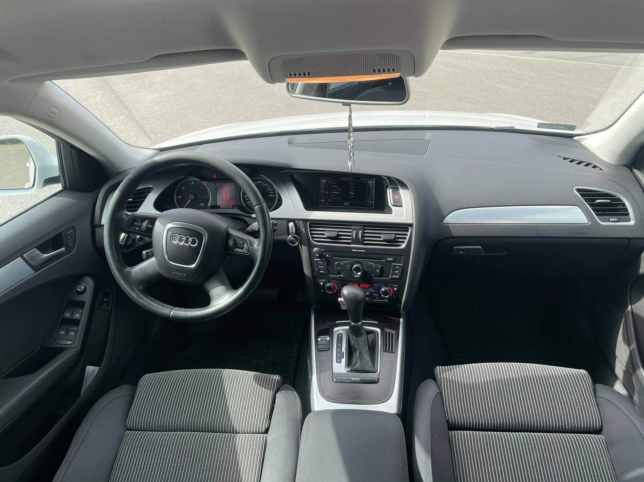 Wnętrze samochodu Audi A4 z widocznymi szarymi fotelami w paski, deską rozdzielczą z srebrnymi akcentami i automatyczną skrzynią biegów, uchwycone z perspektywy przedniego siedzenia.