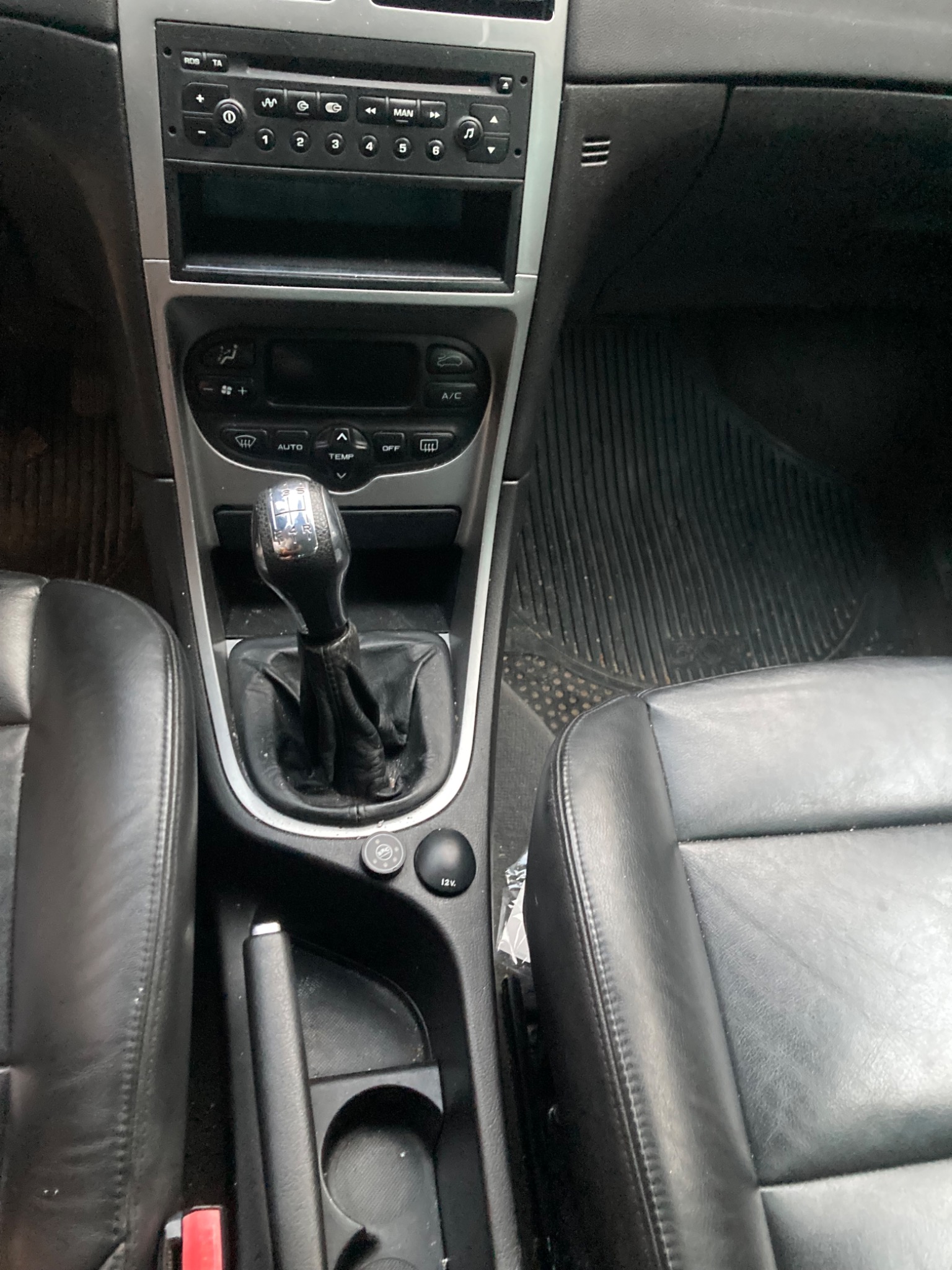 Wnętrze samochodu z widocznymi czarnymi skórzanymi fotelami, dźwignią zmiany biegów, radiem samochodowym oraz gumowymi dywanikami podłogowymi.