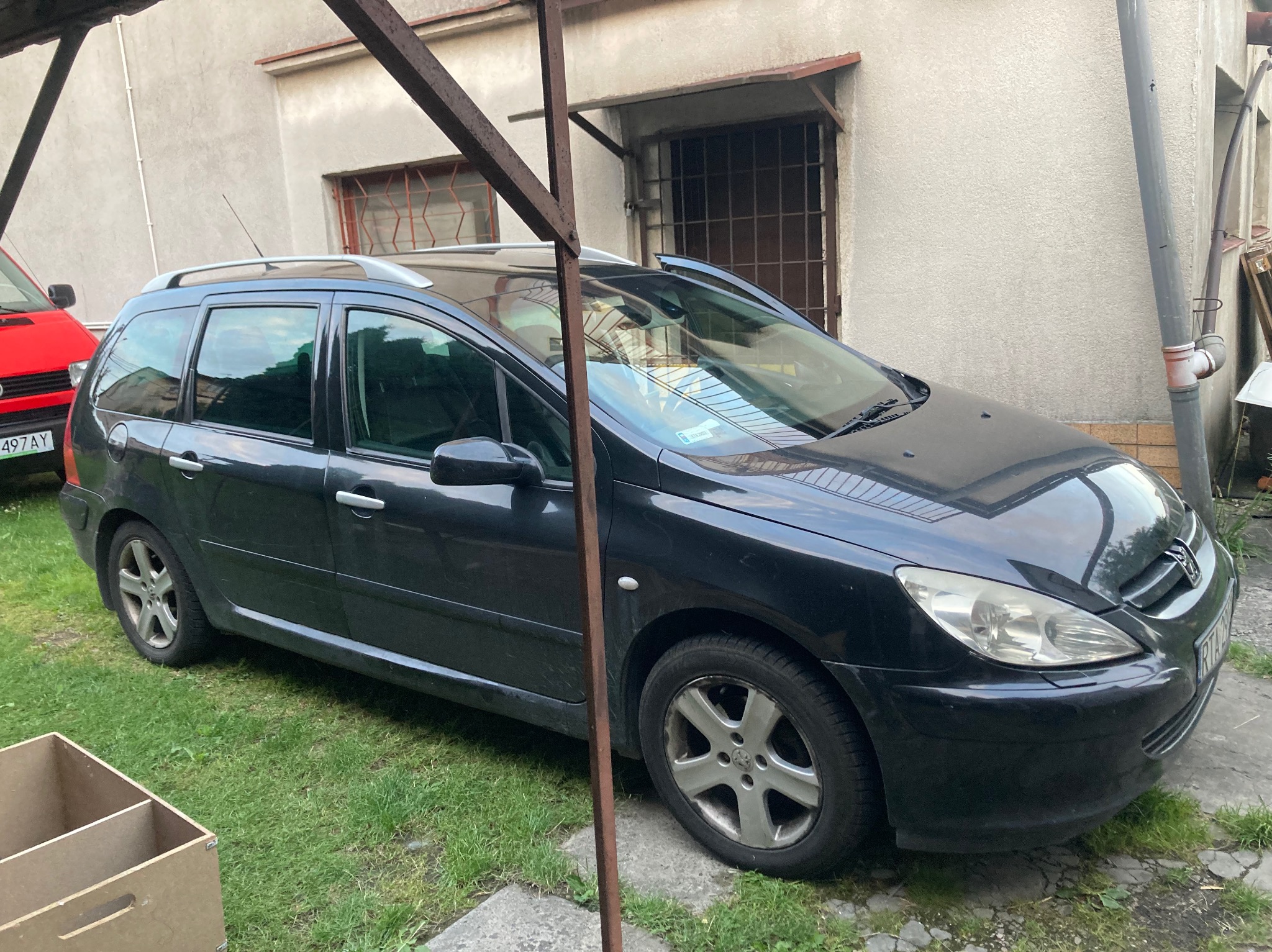 Czarny samochód kombi marki Peugeot zaparkowany na trawie przed budynkiem z kratami w oknach.