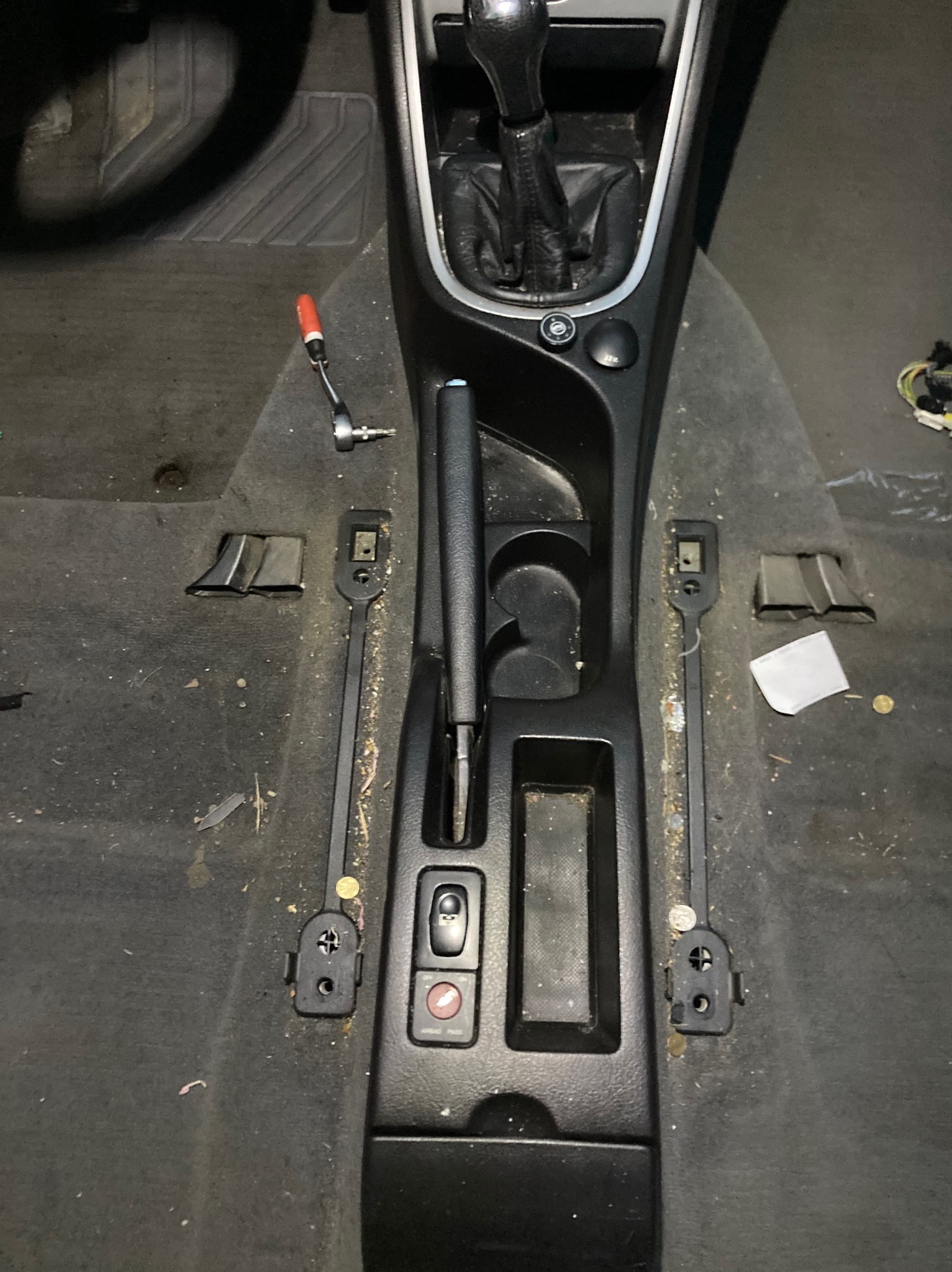 Wnętrze samochodu z widocznym tunelem środkowym, dźwignią zmiany biegów i hamulcem ręcznym, brudna podłoga z widocznymi śladami użytkowania i drobnymi przedmiotami, narzędzie leży na dywaniku.