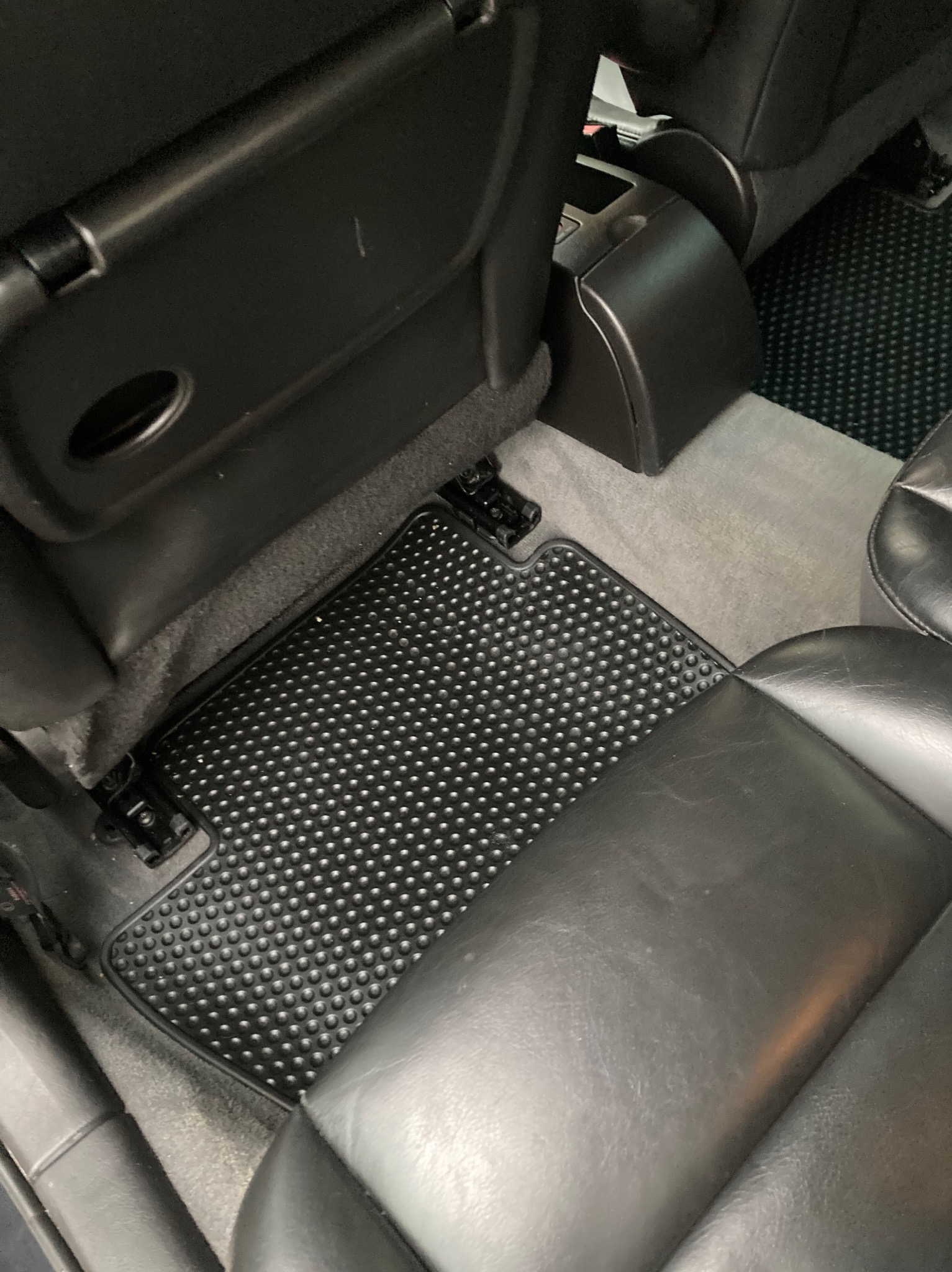 Wnętrze samochodu z czarnym gumowym dywanikiem pod nogami, widoczne szare dywanowe wykończenie pod dywanikiem oraz fragment czarnego skórzanego siedzenia.