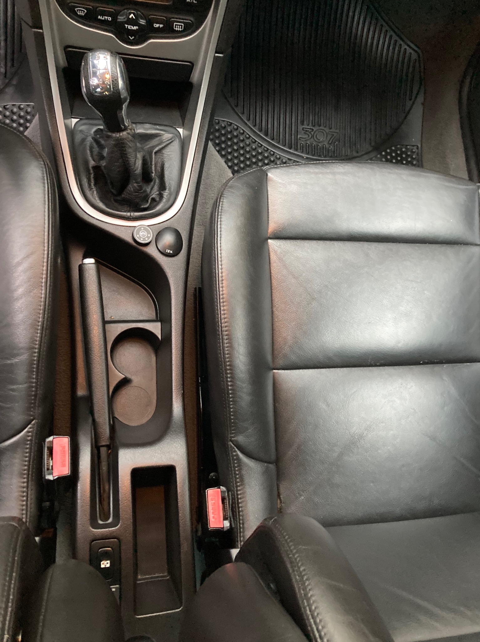 Wnętrze samochodu Peugeot 307: czarna skórzana tapicerka fotela kierowcy z widocznymi śladami użytkowania, gumowy dywanik z logo modelu, dźwignia zmiany biegów i konsola środkowa z uchwytami...