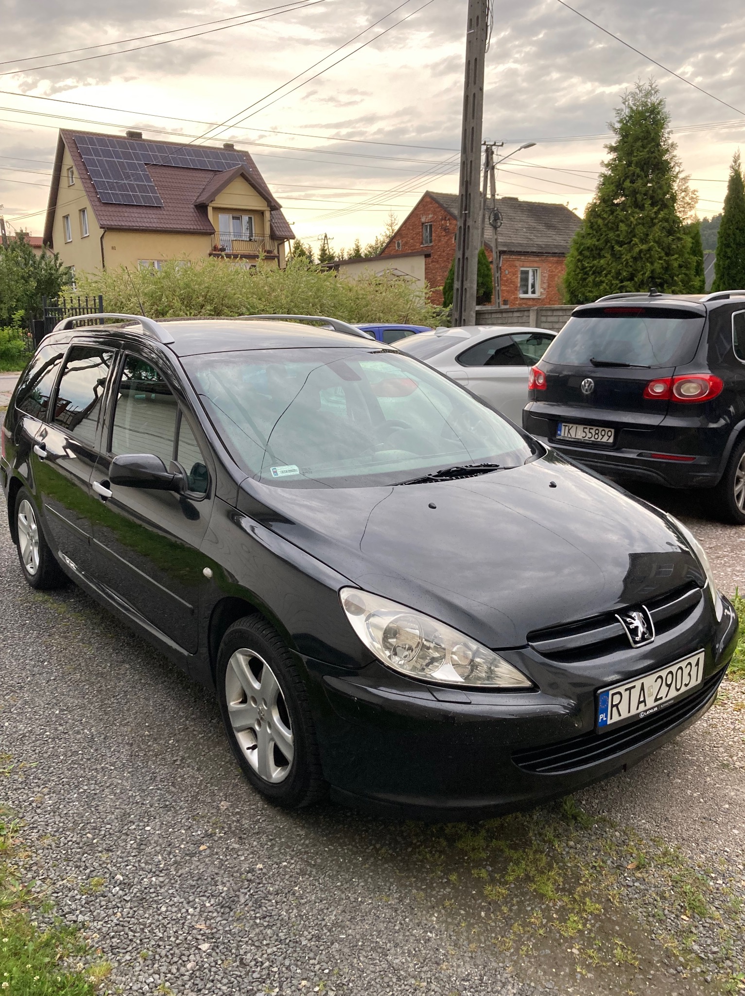 Czarny Peugeot 307 kombi zaparkowany na żwirowym podjeździe, z widocznymi numerami rejestracyjnymi i domami w tle, uchwycony w pochmurny dzień.