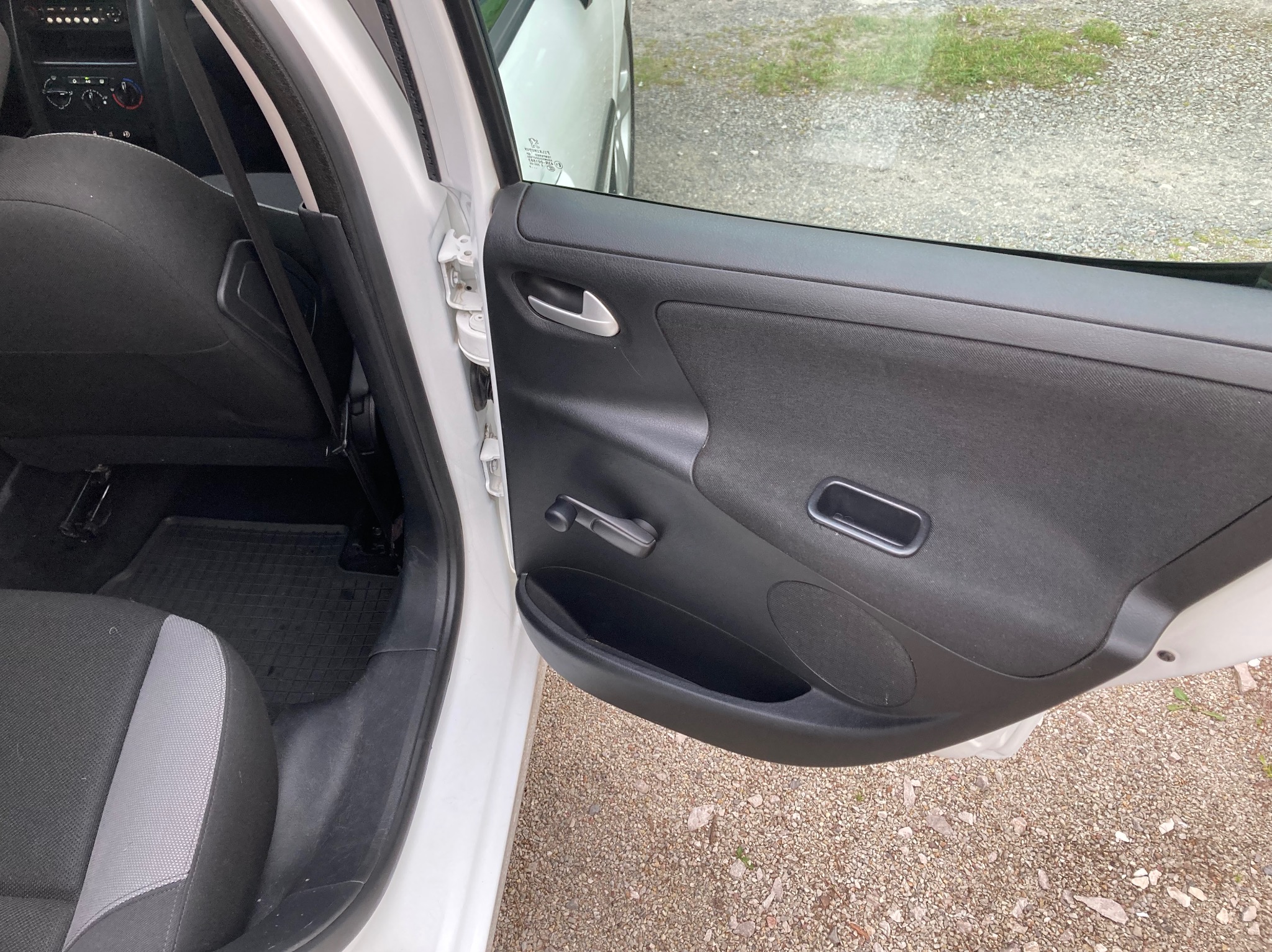 Otwarta biała drzwi samochodu z widocznym wnętrzem: czarna tapicerka, szaro-czarne siedzenie i czarna gumowa wycieraczka pod nogami.
