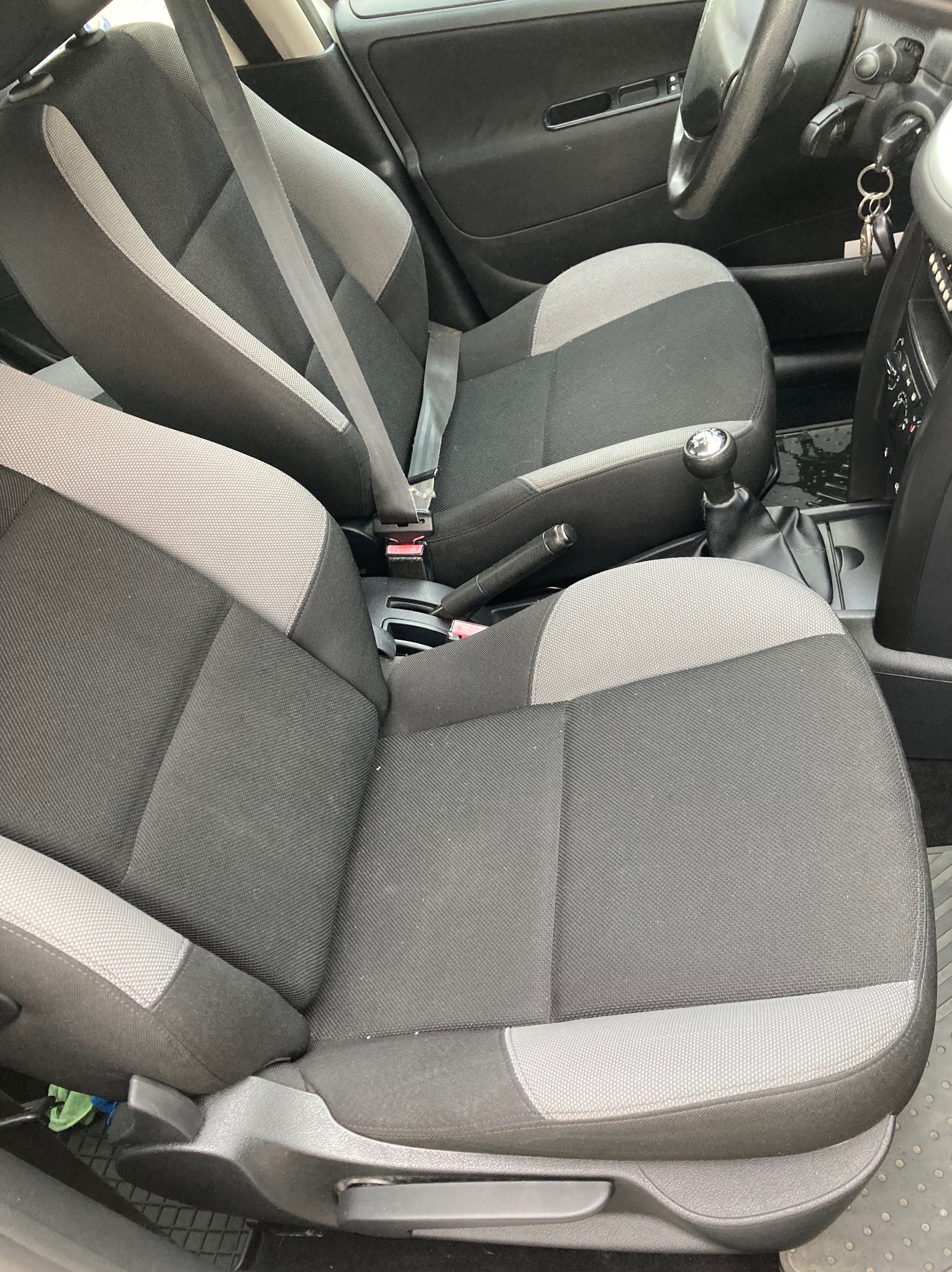 Wnętrze samochodu z widocznymi przednimi fotelami w odcieniach szarości i czerni, z zapiętym pasem bezpieczeństwa na fotelu pasażera i kluczykami w stacyjce, ukazujące stan przed czyszczeniem.