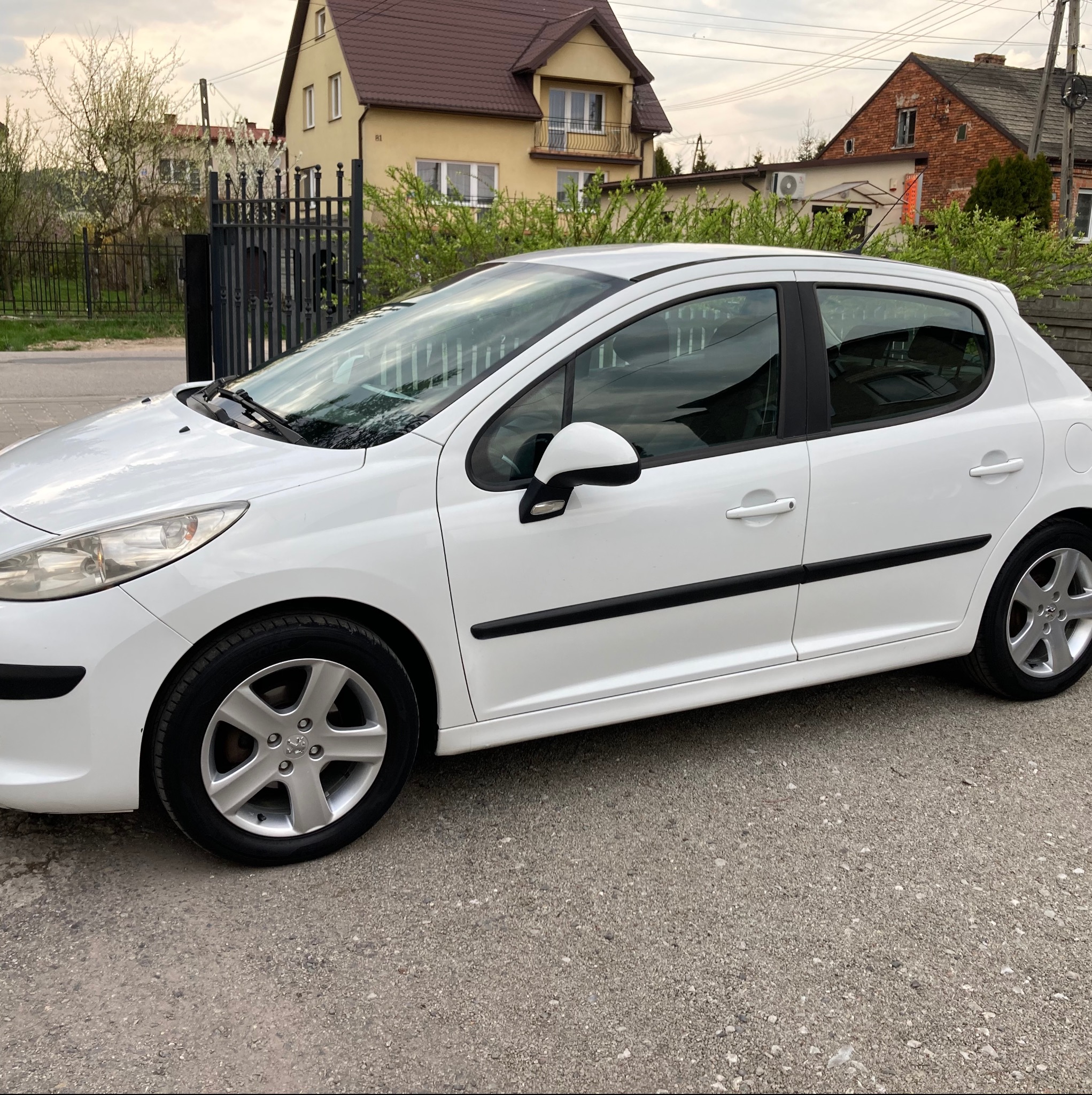 Biały Peugeot 207 zaparkowany na szarym asfalcie, z widocznymi domami w tle i zielenią, prezentowany z boku w pochmurny dzień.