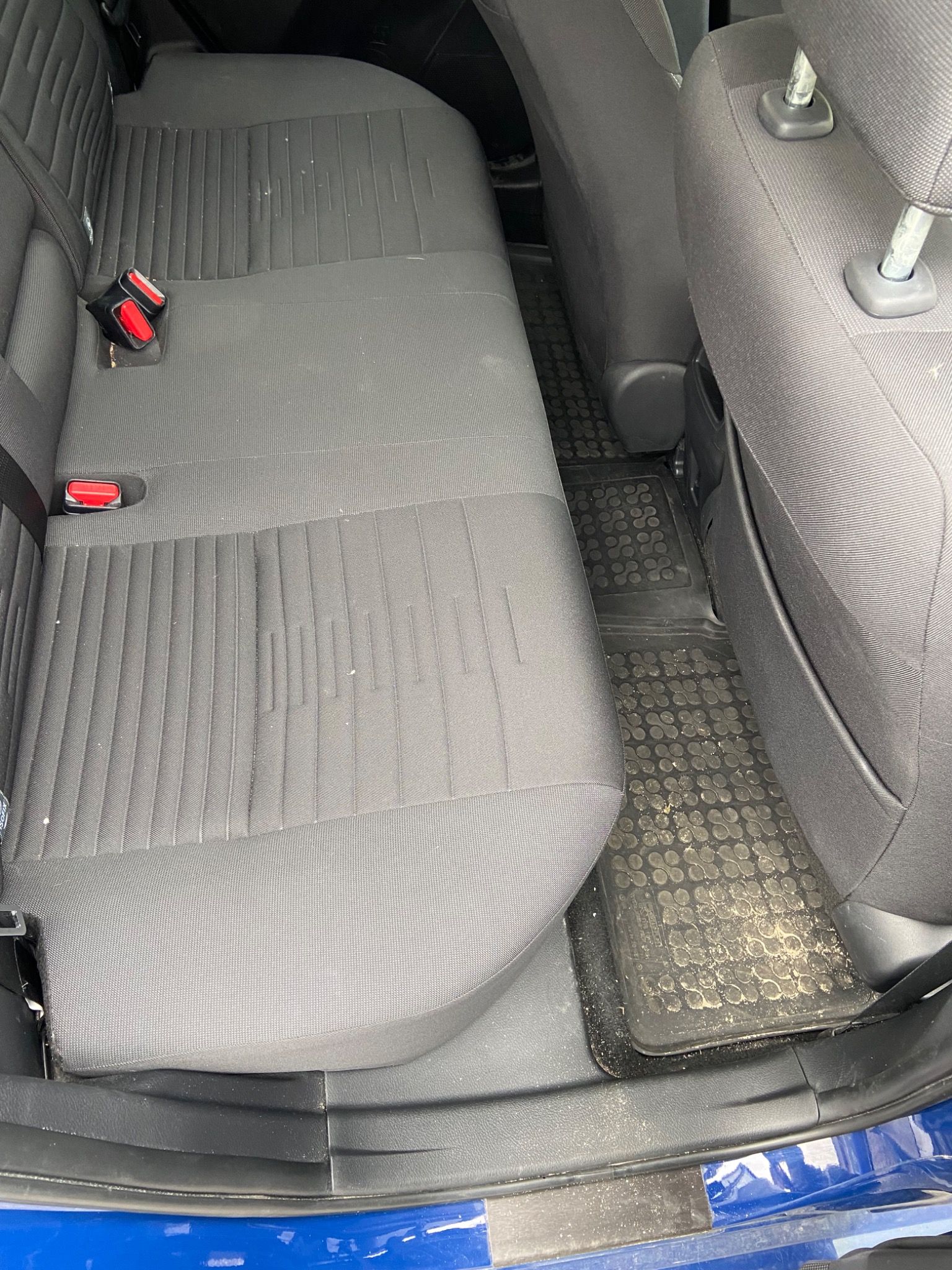 Brudna tylna kanapa samochodu z poplamionymi dywanikami gumowymi, widoczne plamy i okruchy na szarej tapicerce.