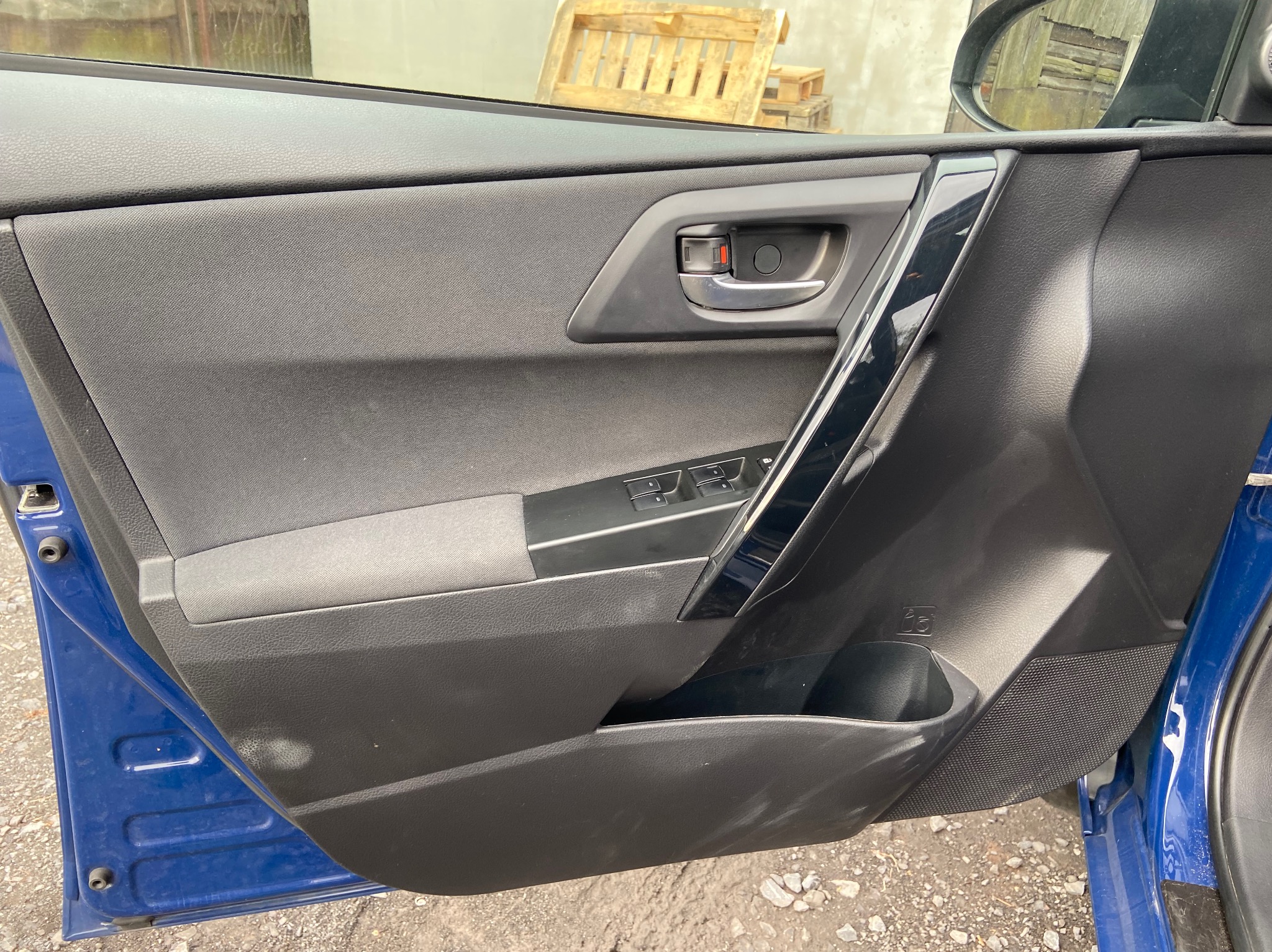Wnętrze samochodu: tapicerka drzwi w kolorze czarnym i szarym, widoczne elementy sterowania szybami, klamka z czerwonym akcentem i kieszeń na drobiazgi, zarys niebieskiego nadwozia.
