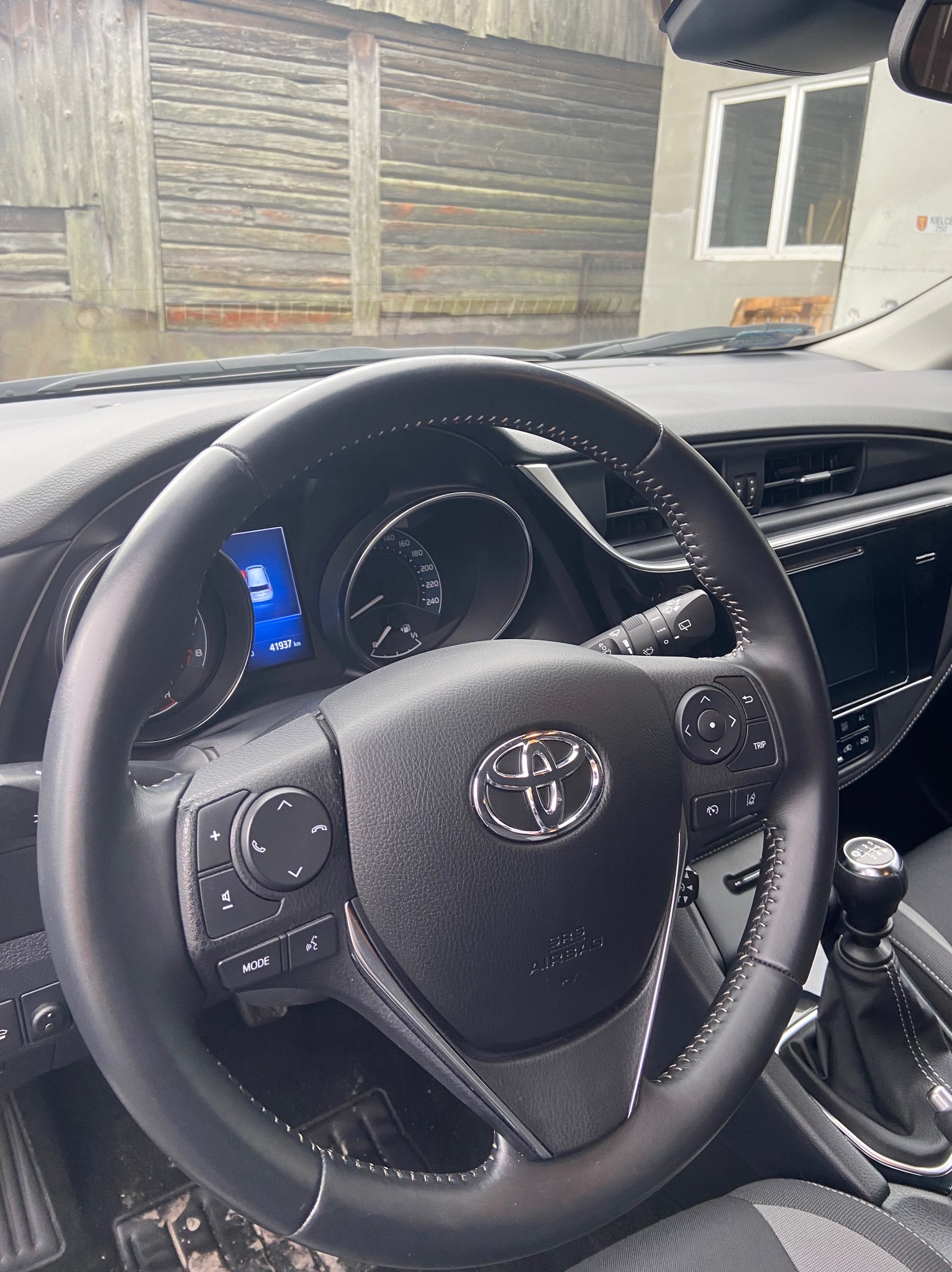 Wnętrze samochodu Toyota z widocznym kokpitem, kierownicą z logo, deską rozdzielczą i skrzynią biegów na tle drewnianej ściany budynku.