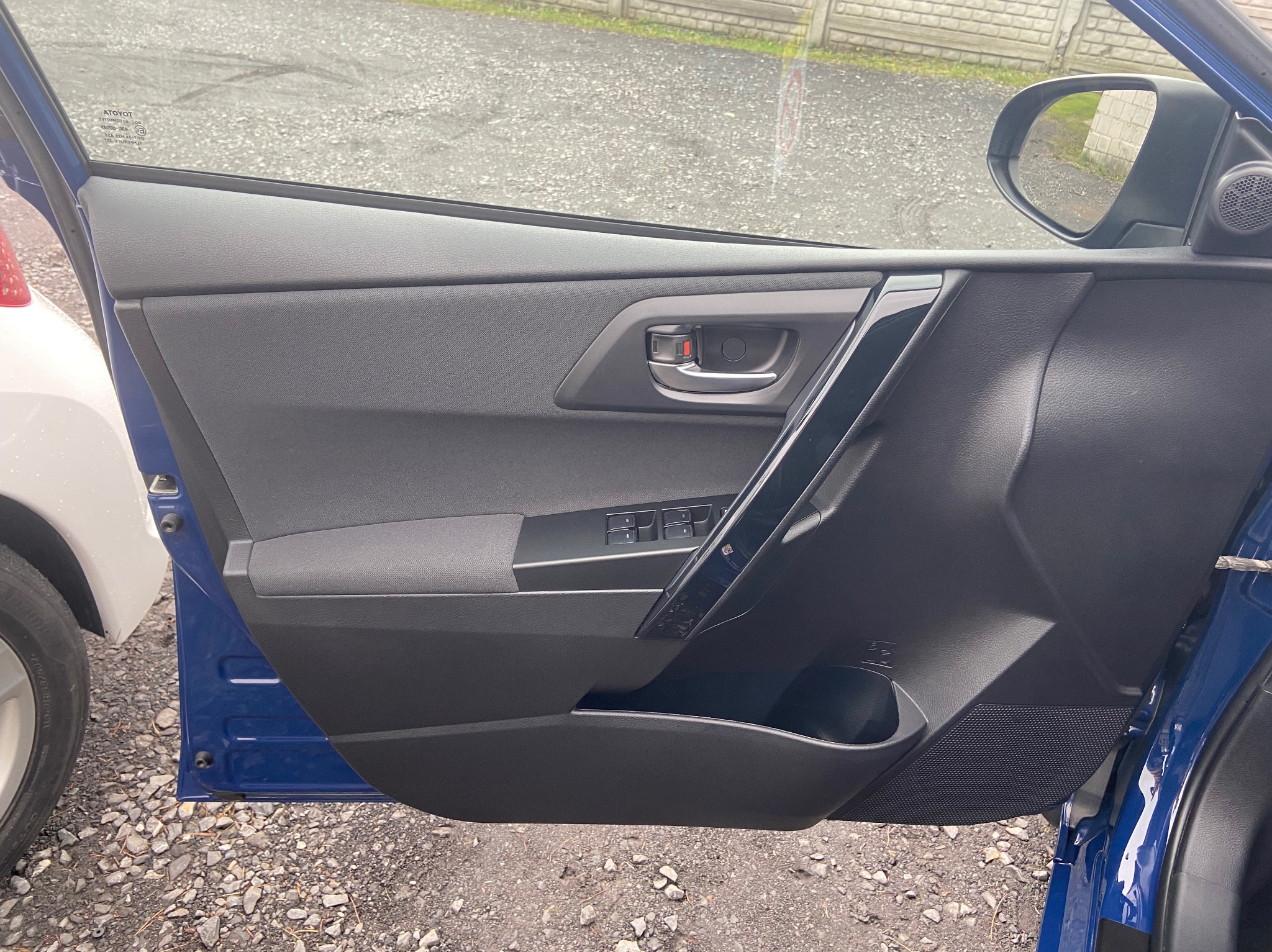 Wnętrze błękitnego samochodu Toyota, widoczny panel drzwi z elementami tapicerowanymi i plastikowymi, uchwyt, przyciski sterowania szybami oraz kieszeń, na zewnątrz widoczny fragment innego auta...