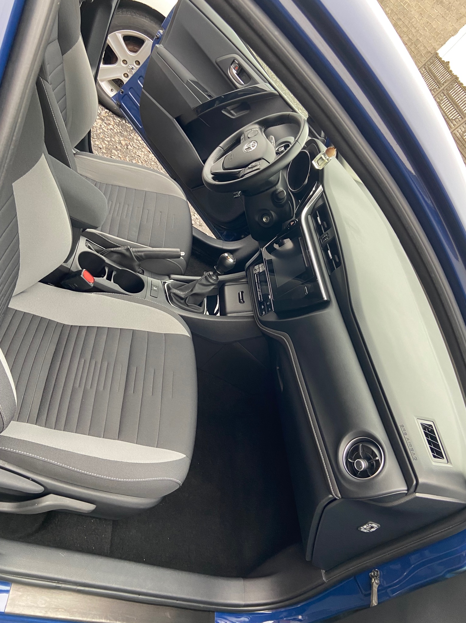 Wnętrze niebieskiego samochodu z szaro-białą tapicerką, widok z otwartymi drzwiami na deskę rozdzielczą, kierownicę i fotele.