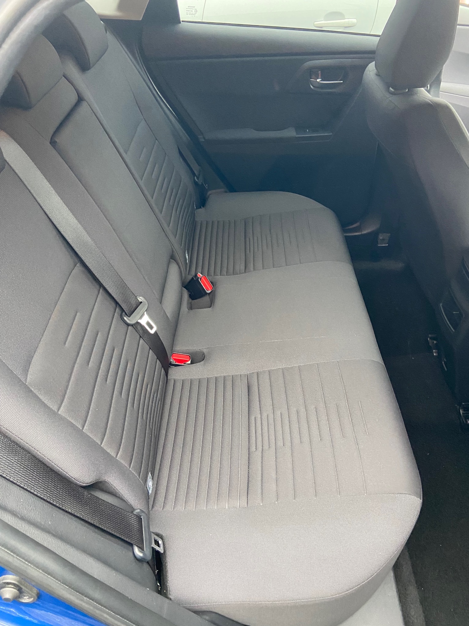 Szare tylne siedzenie samochodu z widocznymi pasami bezpieczeństwa i zagłówkami, z delikatnym wzorem w pionowe prążki na tapicerce.